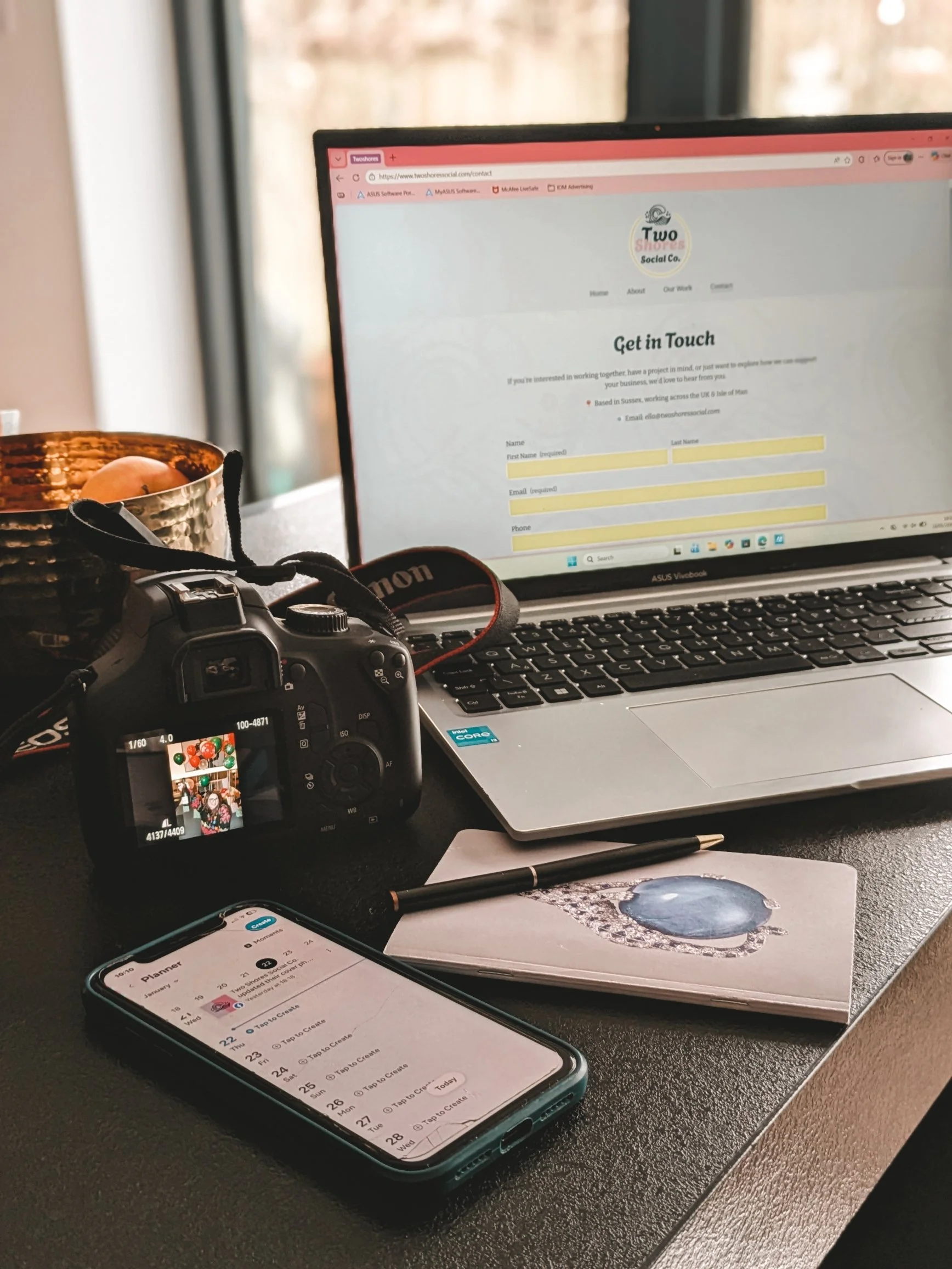 A desk setup with a laptop displaying a contact form, a digital camera showing a photo on its screen, a smartphone with a calendar app, a pen, a small notebook with a water droplet illustration, and a copper mug, near a window with natural light.