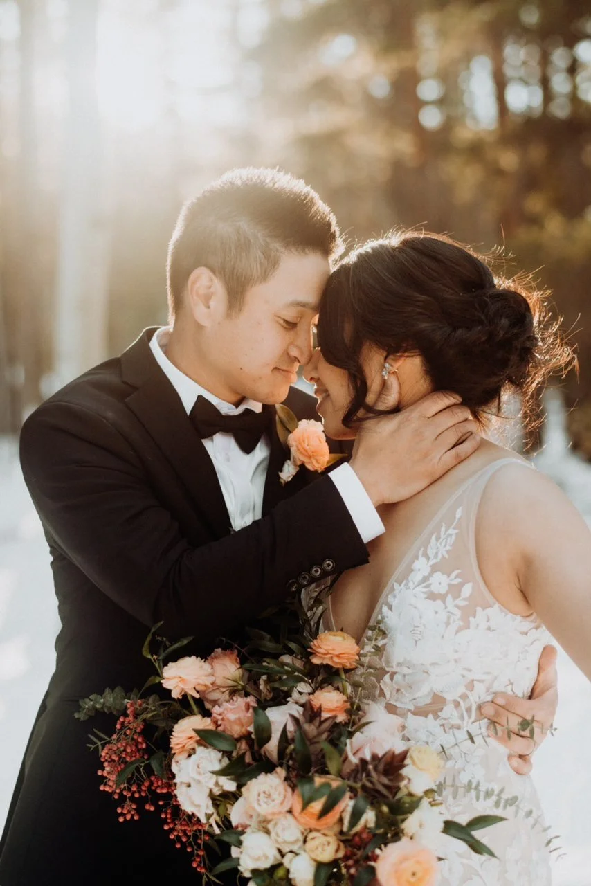 A bride and groom share a close, intimate moment outdoors, with the groom gently holding the bride's face, both smiling with their foreheads touching. The groom wears a black tuxedo with a bow tie and a boutonniere, and the bride is in a lace wedding gown, holding a large bouquet of peach, white, and pink flowers.