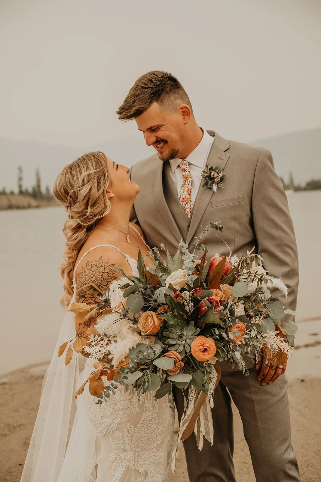 A bride and groom standing on a beach, smiling at each other, with the bride holding a large bouquet of flowers and greenery.