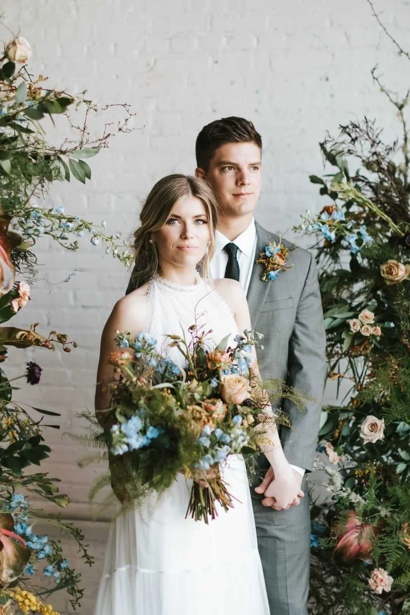 A bride and groom pose for a wedding photo surrounded by floral arrangements.