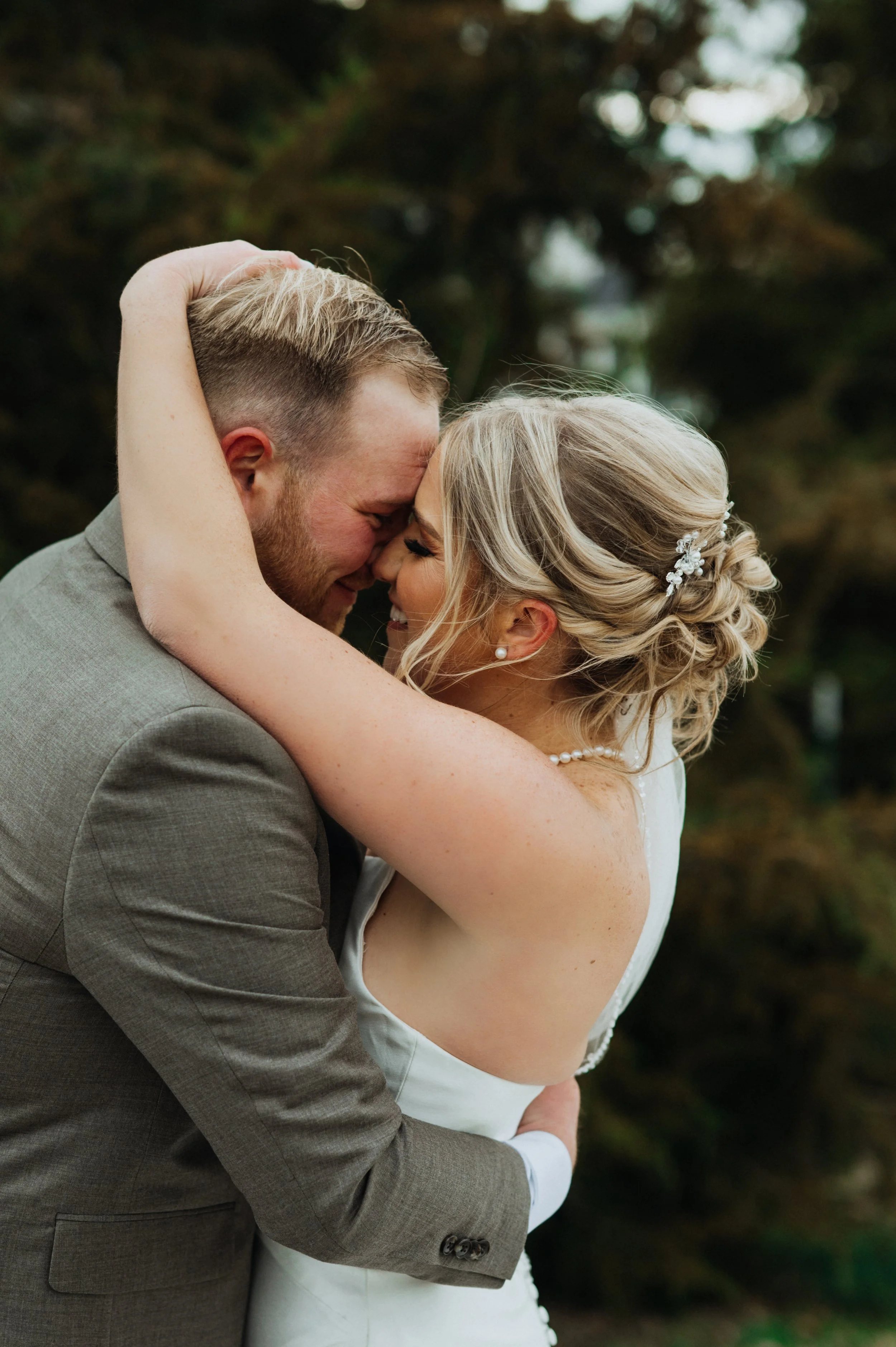 A happy couple on their wedding day embracing outdoors, with their foreheads and noses touching and smiling.