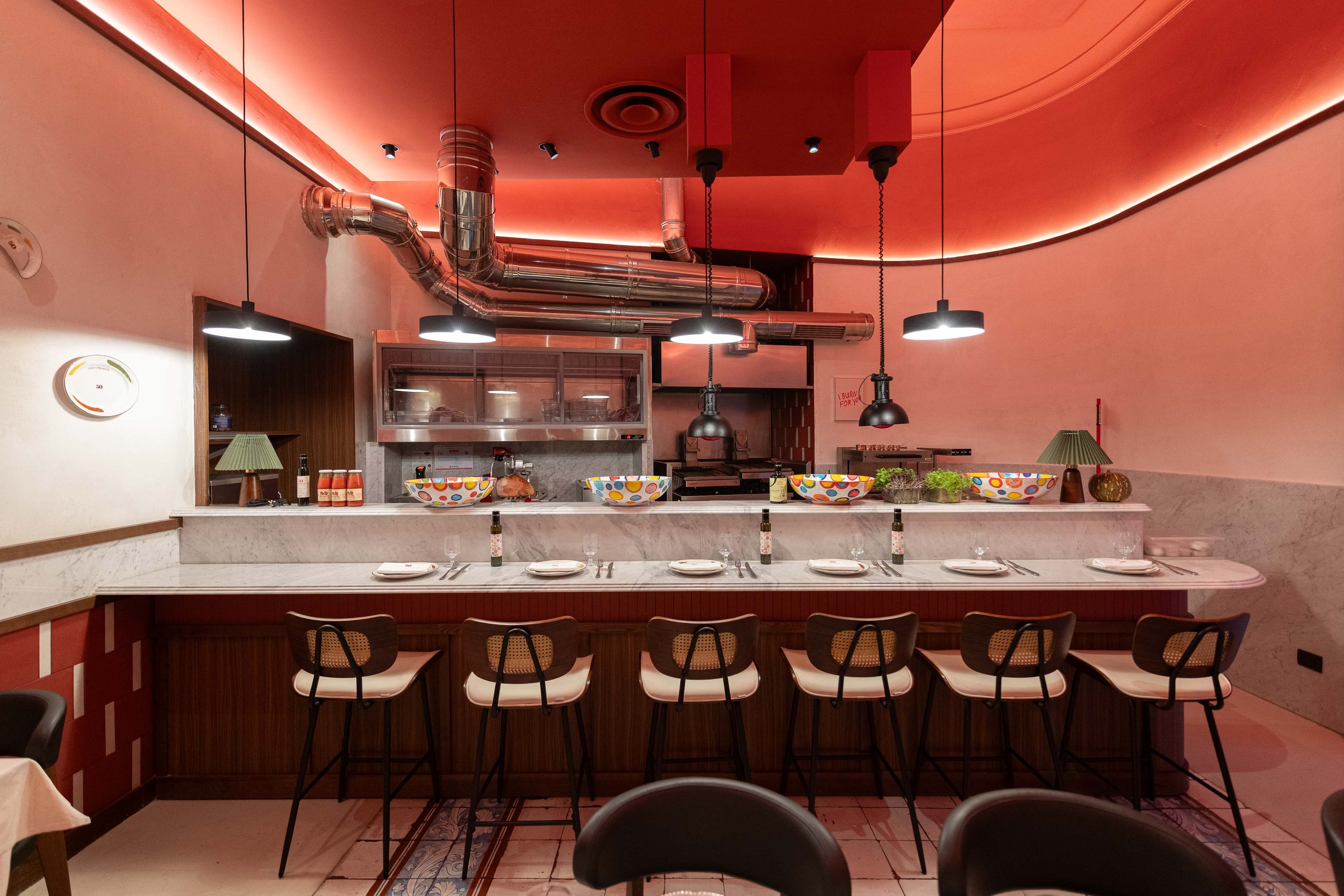 Restaurant interior with a bar seating area, colorful bowls, table settings, and kitchen in the background with industrial pipes and pendant lighting.