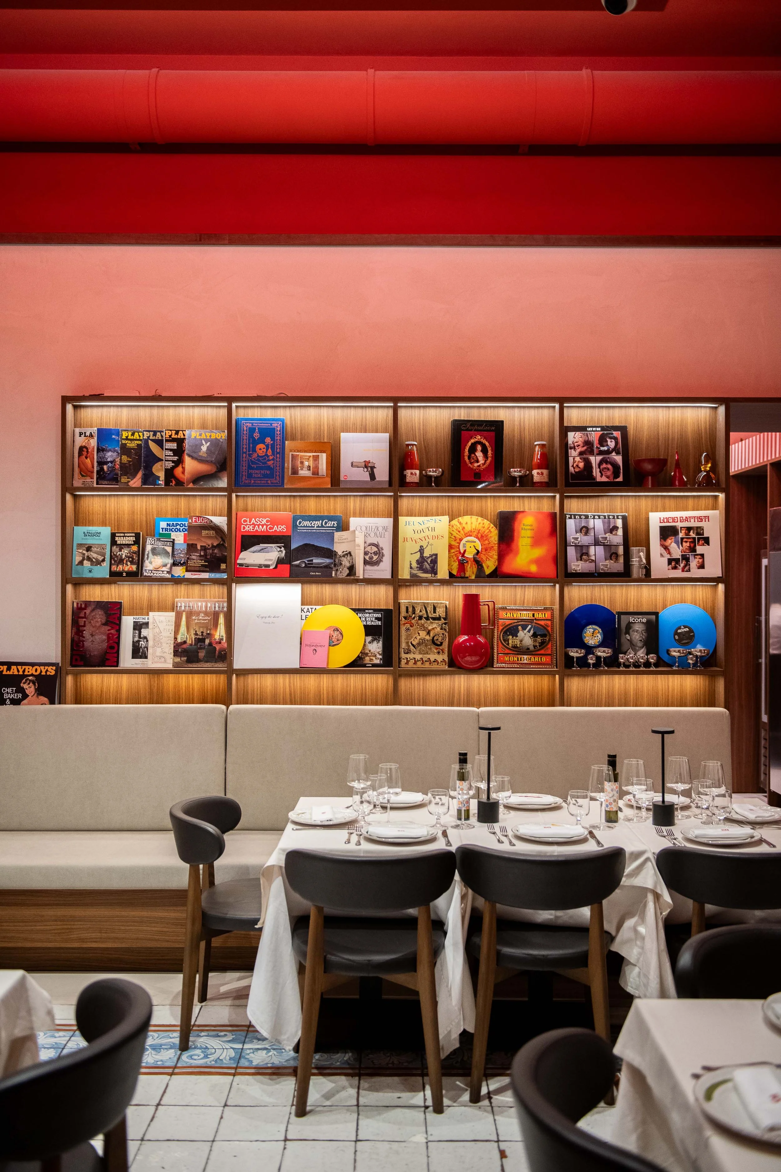 A restaurant interior with a wooden shelving unit filled with books, magazines, decorative items, and records, a long beige upholstered bench, and a table set for dining with white tablecloths, glassware, and place settings.