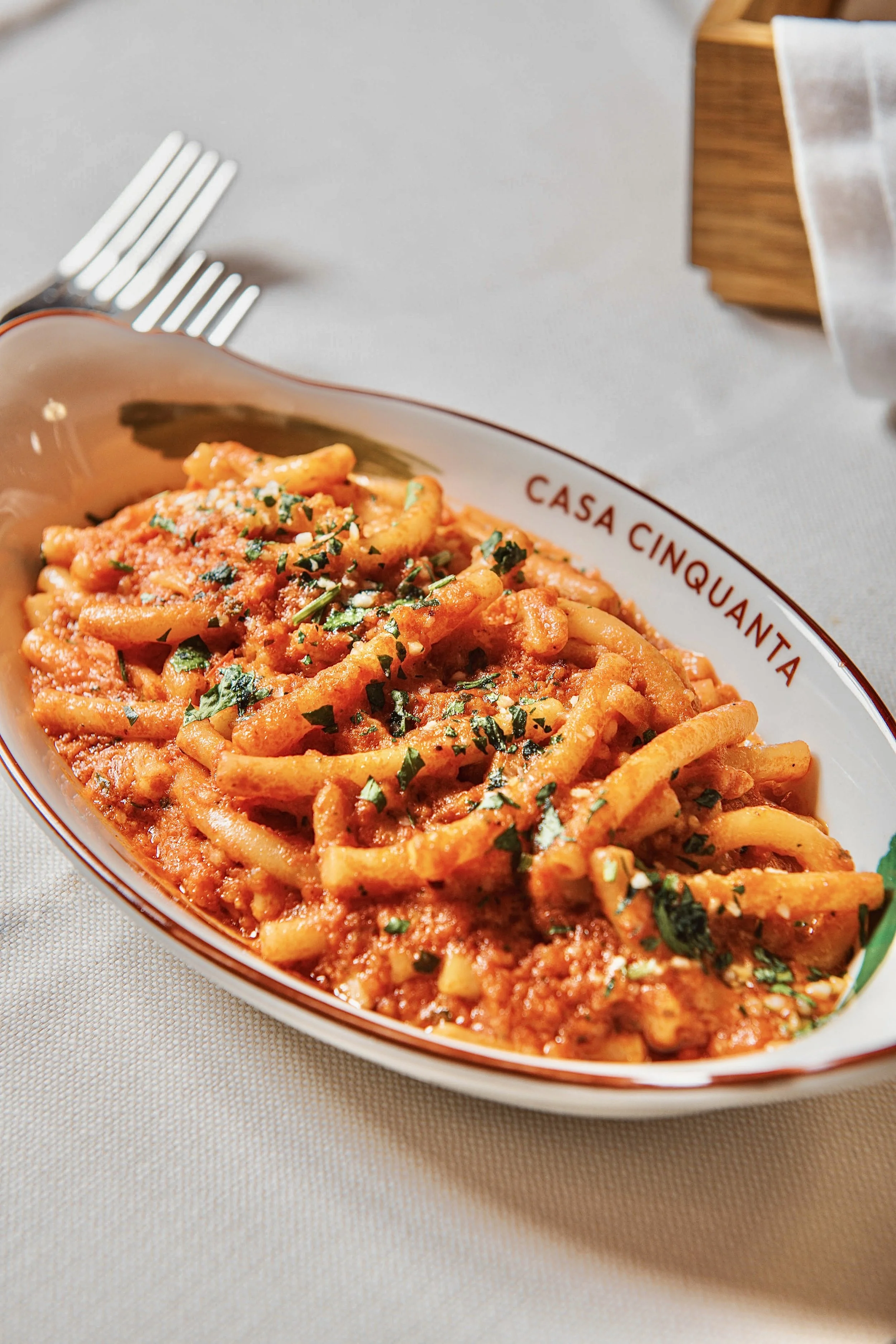 A dish of pasta, likely spaghetti, topped with tomato sauce and chopped herbs, served in a white bowl with red lettering that reads 'Casa Cinquanta' on a beige tablecloth.