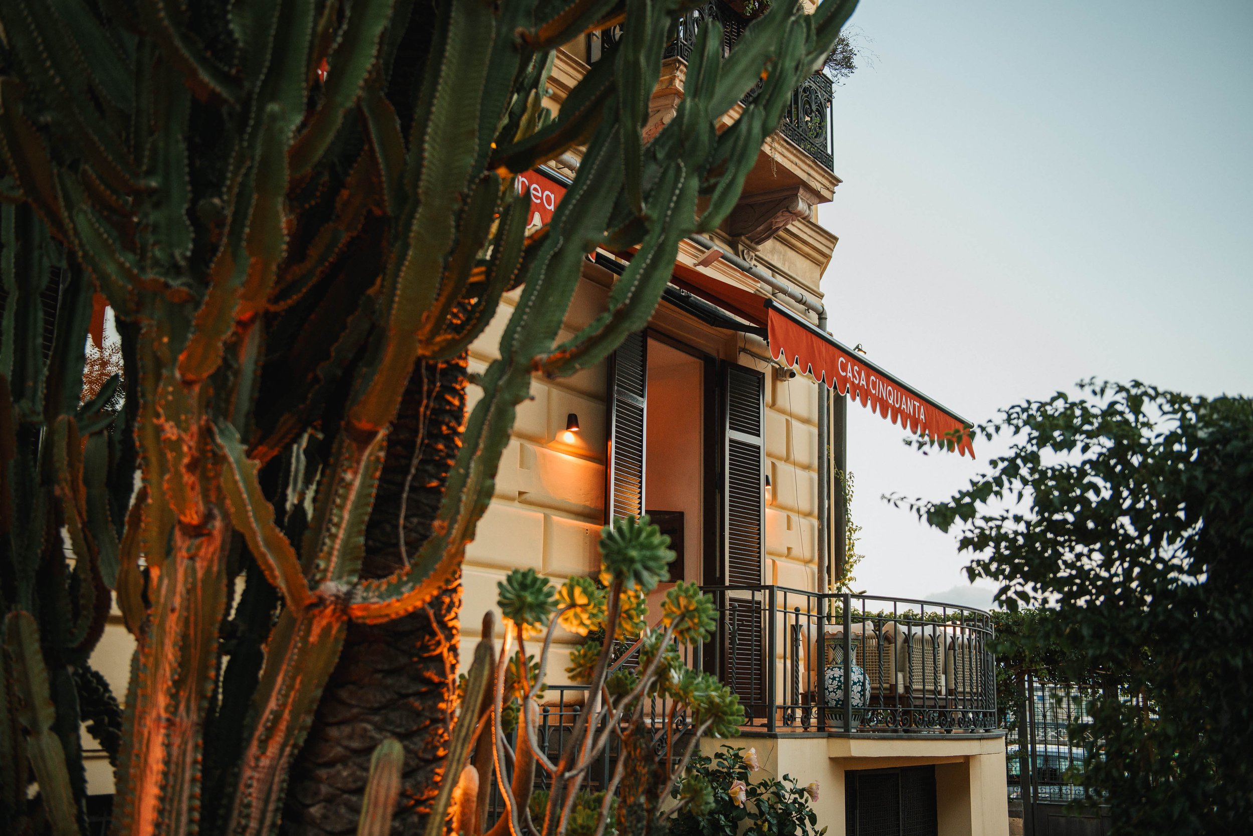 A building with a balcony has an orange awning with the words Casa Cinquanta, surrounded by greenery and cacti.