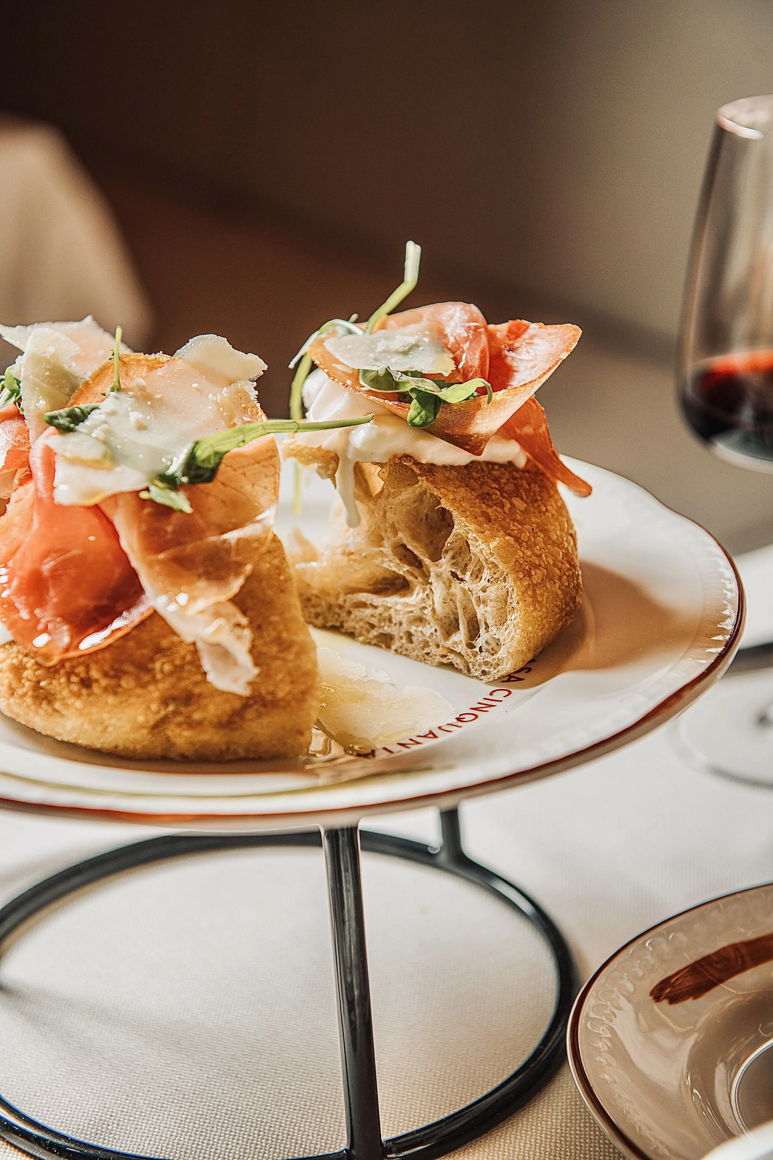 Close-up of an Italian appetizer with prosciutto, fresh greens, shaved Parmesan cheese, and mozzarella on toasted bread served on a stand with a glass of red wine in the background.