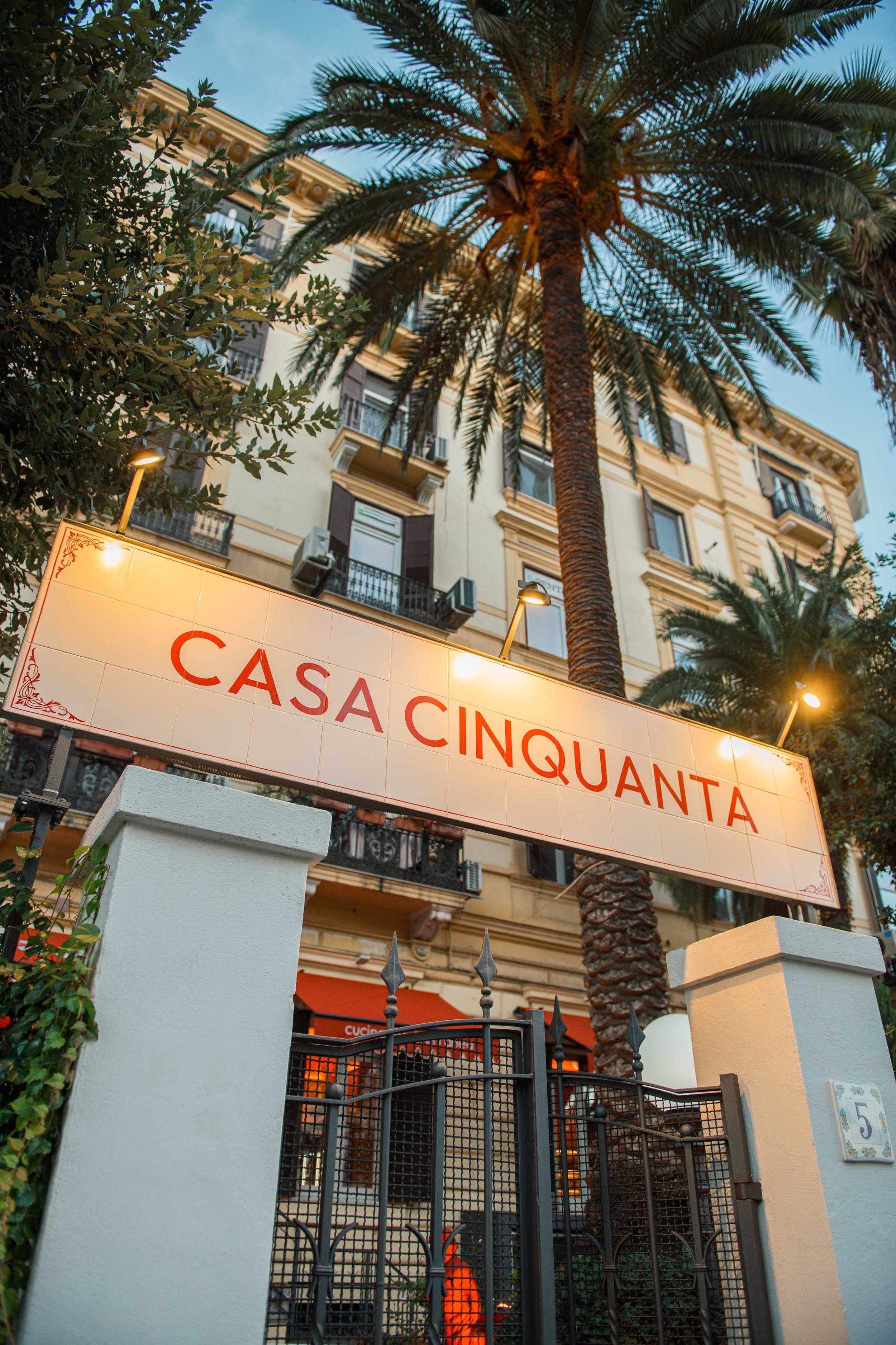 A sign reading 'Casa Cinquanta' in front of a building with palm trees and an outdoor patio area.
