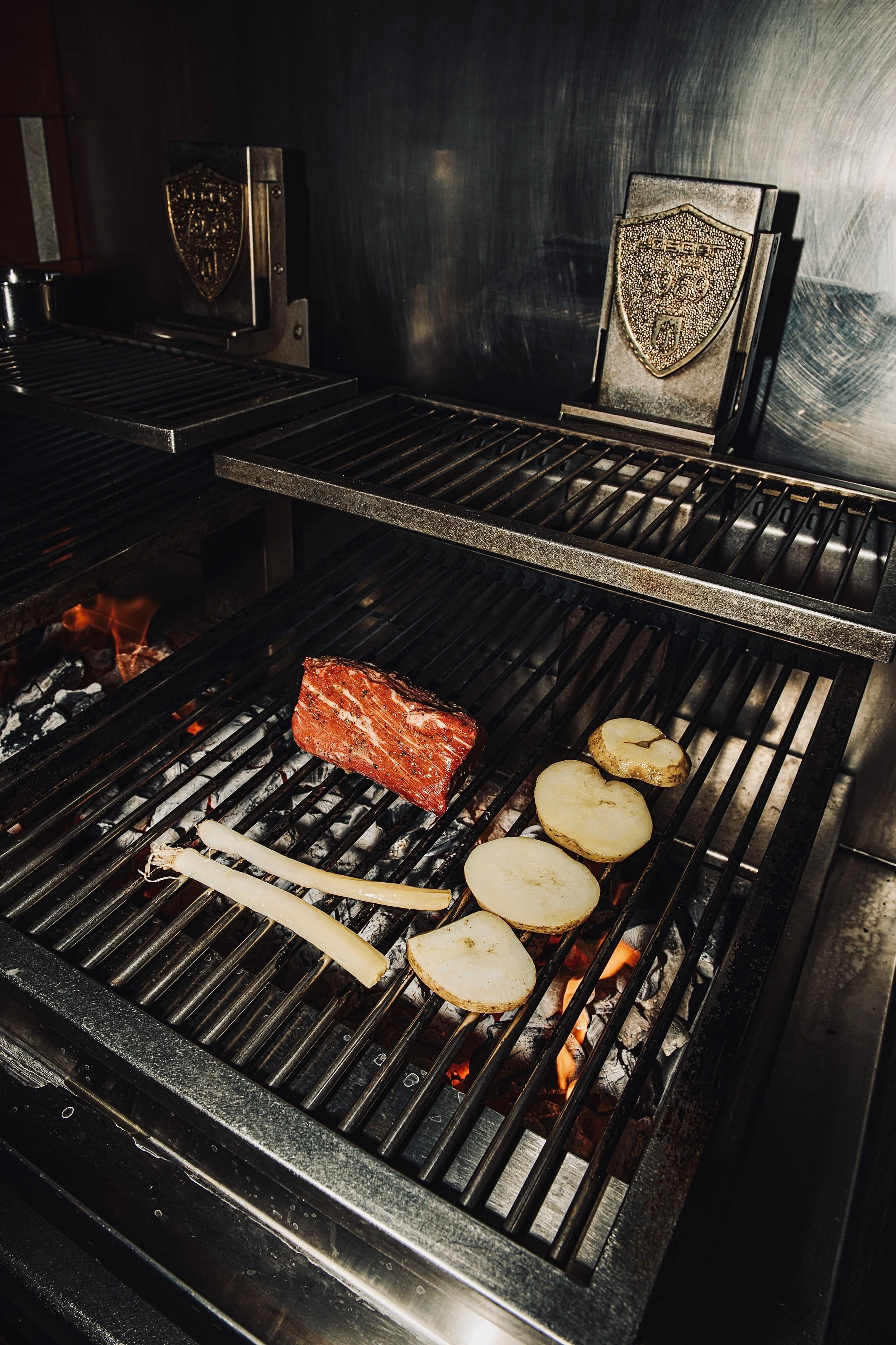 Grill with potato slices, two pieces of white onion, and a piece of red meat cooking over hot coals.