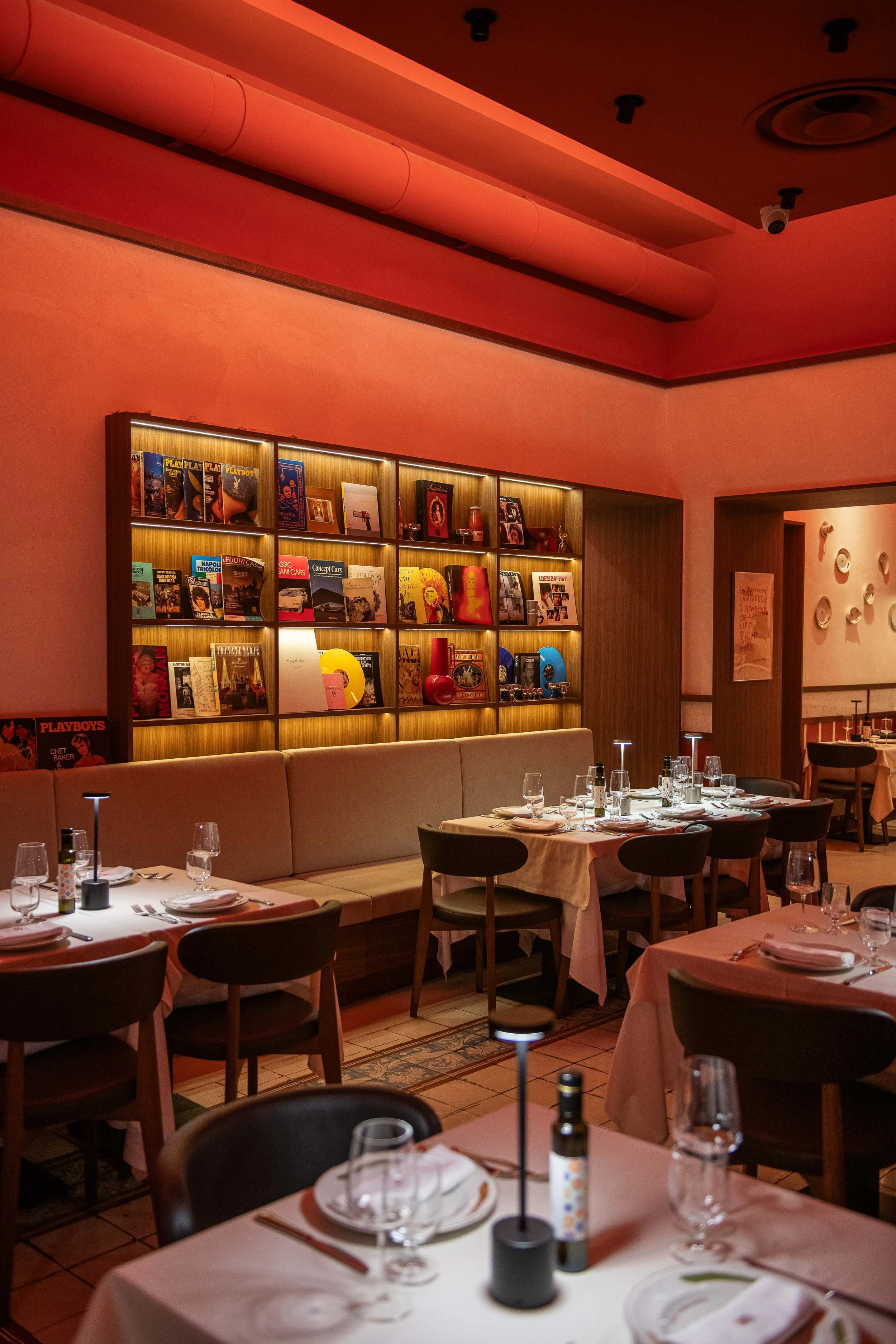 Elegant restaurant interior with tables set with glassware and cutlery, a beige cushioned seating area, and a backlit bookshelf displaying books and decorative objects, illuminated with warm lighting and pinkish ceiling lights.