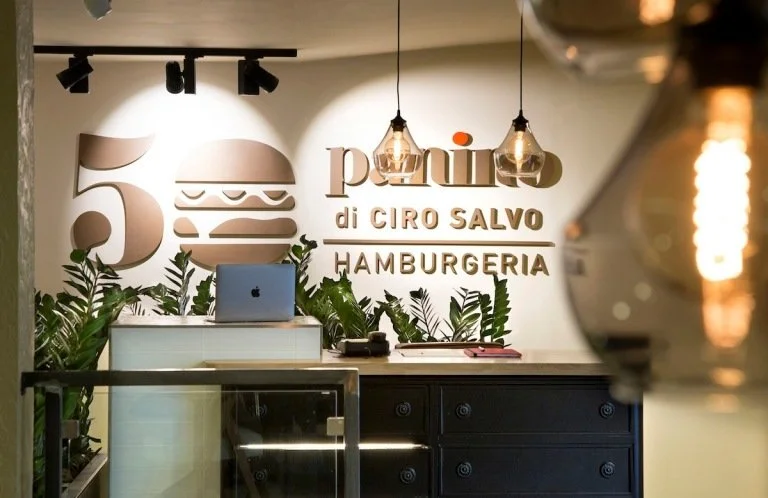 Interior of a burger restaurant with a sign that reads 'panu di GIRO SALVO HAMBURGERIA', green plants, a white counter with a laptop, and hanging Edison-style light bulbs.