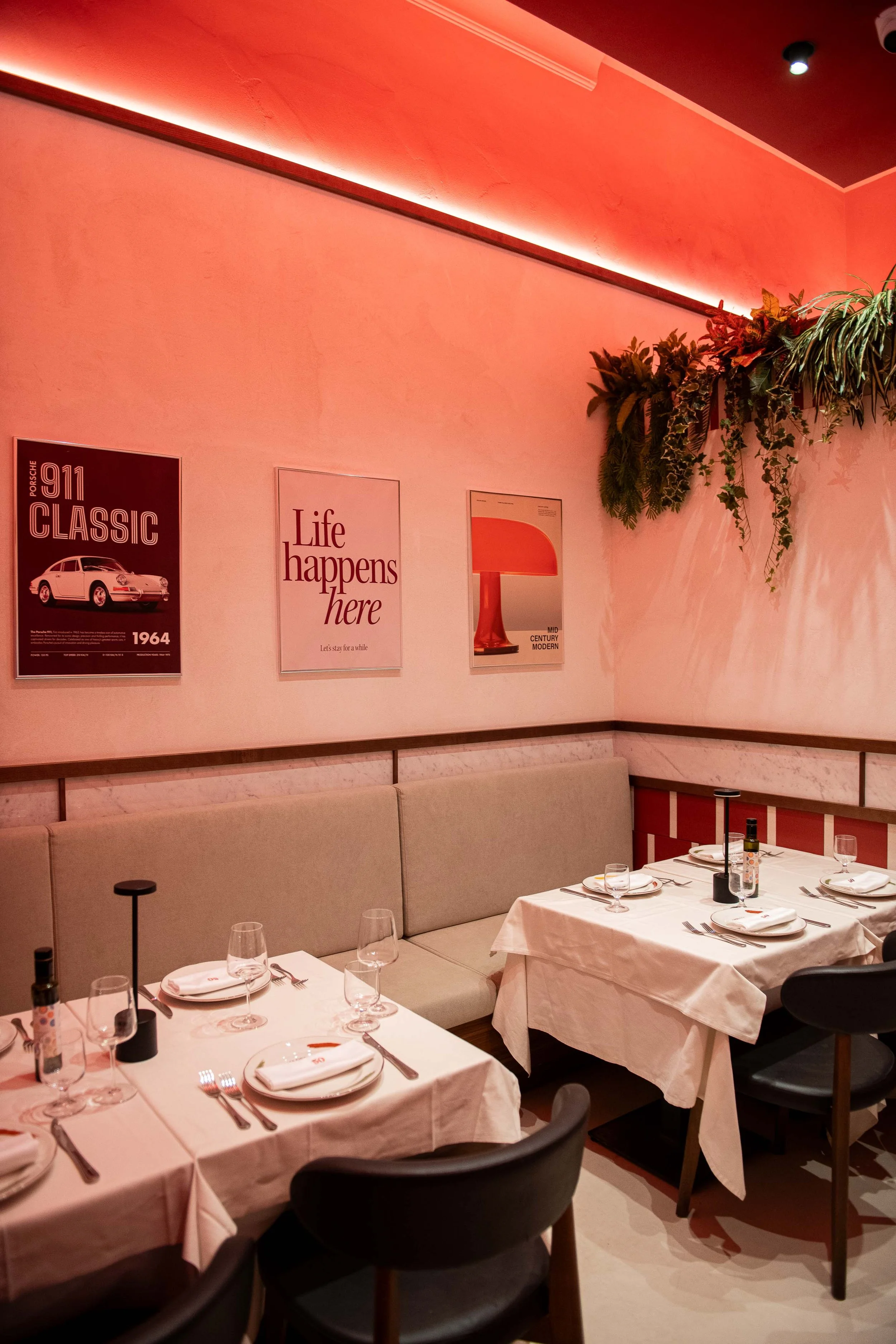 Interior of a restaurant with tables set for dining, pink lighting, framed posters on the wall, and a green plant hanging from the ceiling.
