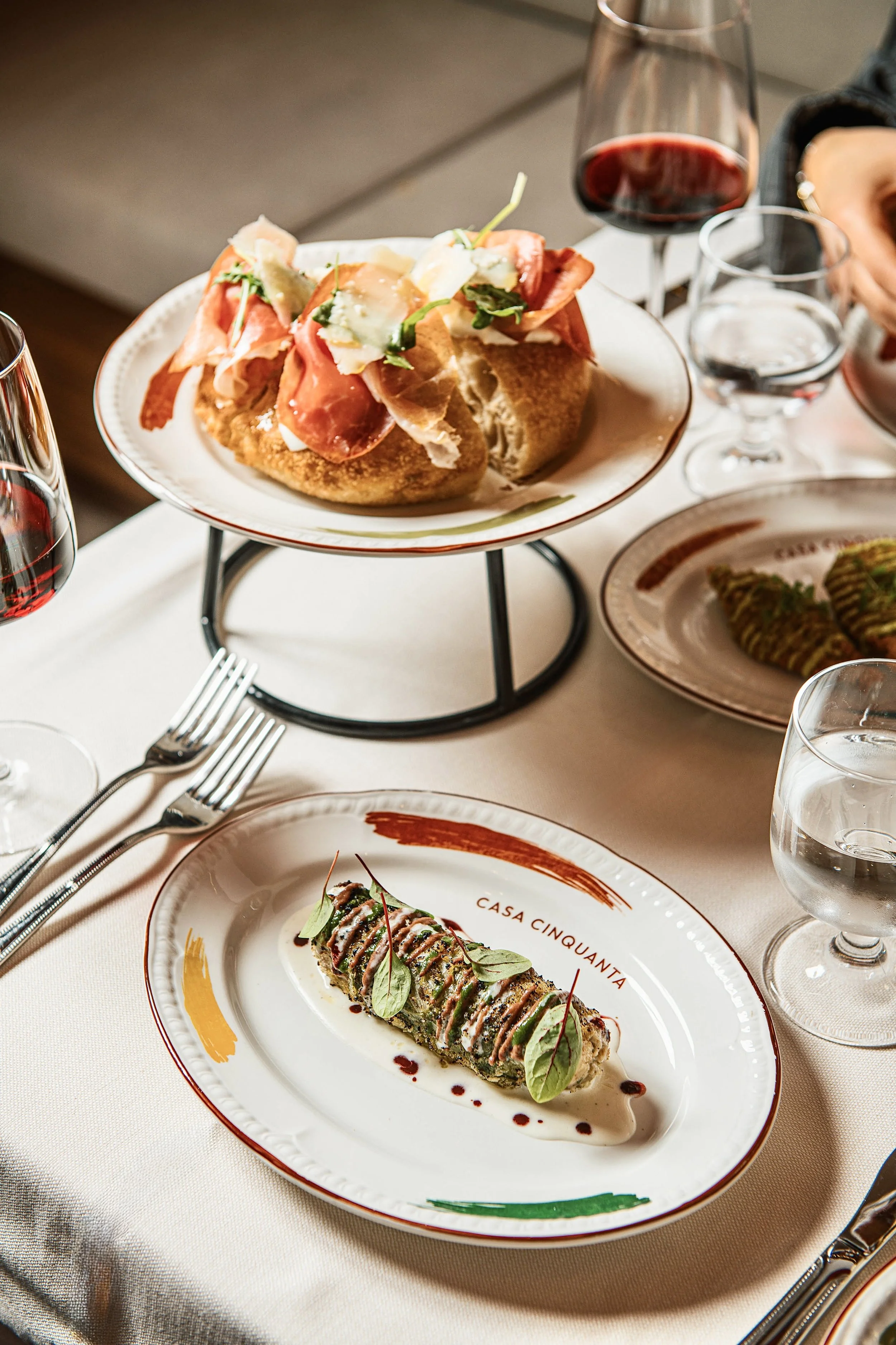 A plated cannelloni dish with sauce and garnishes on a white plate labeled 'Casa Cinquanta', placed on a white tablecloth, with other plates, wine glasses, and cutlery around.