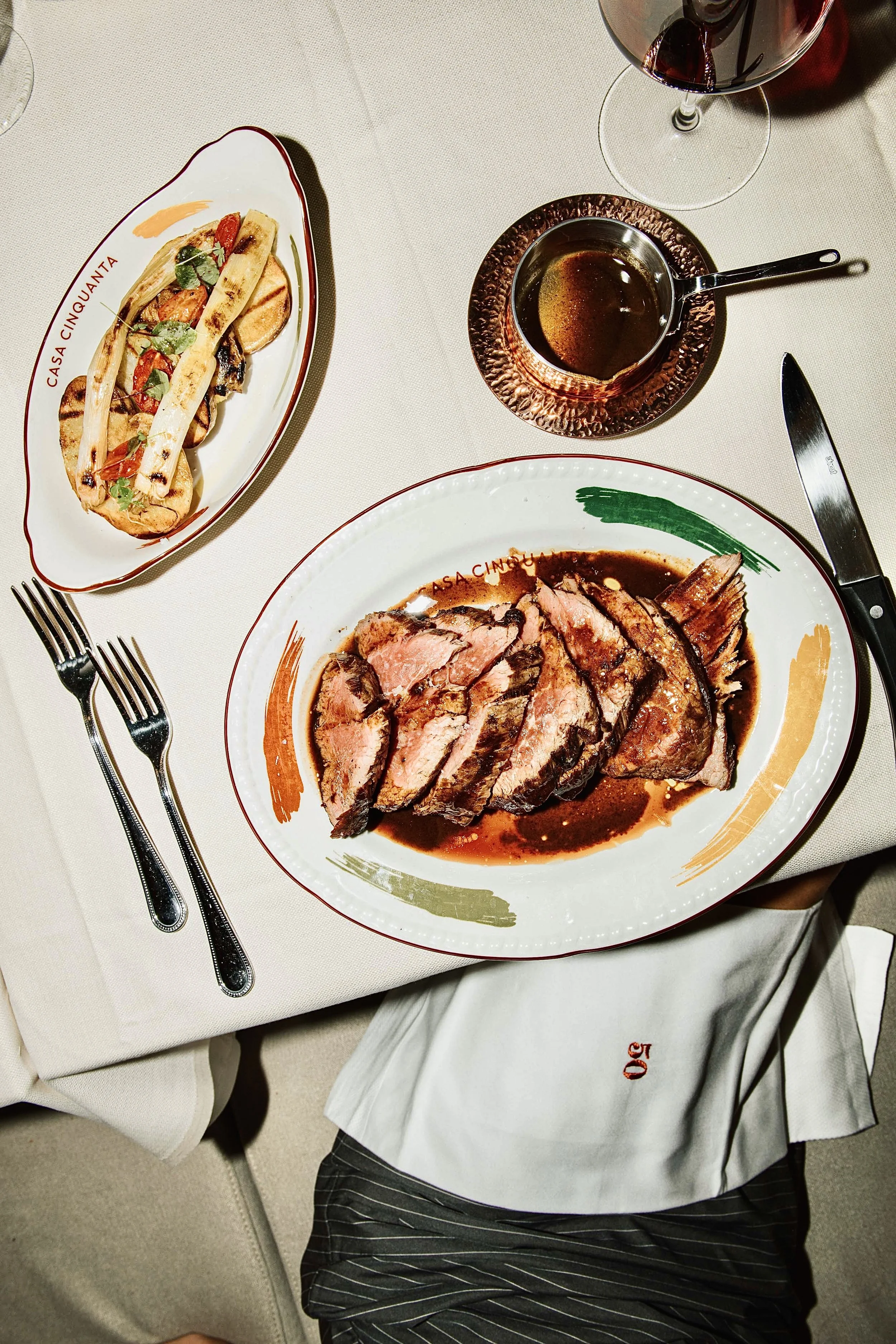 Sliced cooked beef with sauce on a decorative oval plate, grilled bread with toppings on a separate oval dish, glass of red wine, small copper sauce pot with sauce, set on a white tablecloth with cutlery.