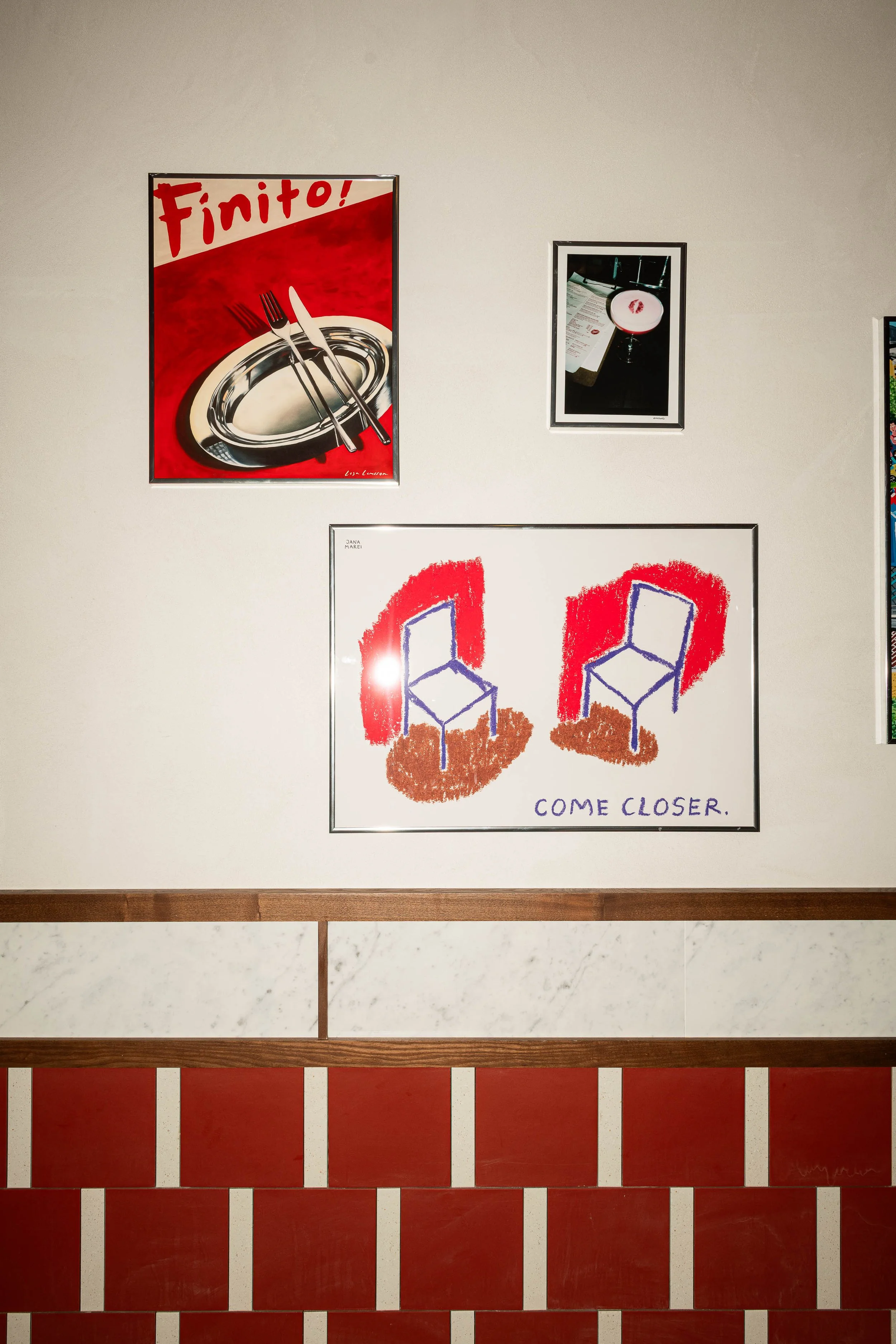 Art display on wall featuring a colorful illustration of two chairs with the text 'COME CLOSER,' a framed photograph of a dining table with a drink and a menu, and a poster with a red background, a white oval plate, and silverware, with the word 'Finito!' in red.