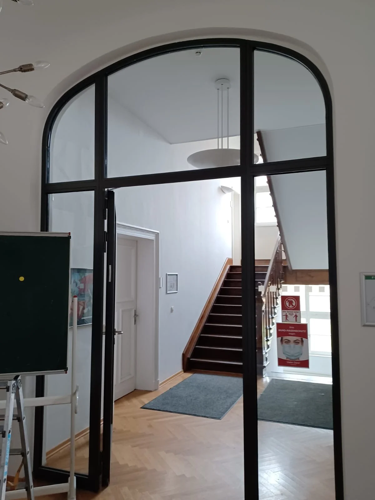 Innenaufnahme eines Eingangsbereichs mit schwarzen Metallrahmen, weißen Wänden, Holztreppe, grauen Teppichen, einem Wandbild und einem Schild mit Maske.