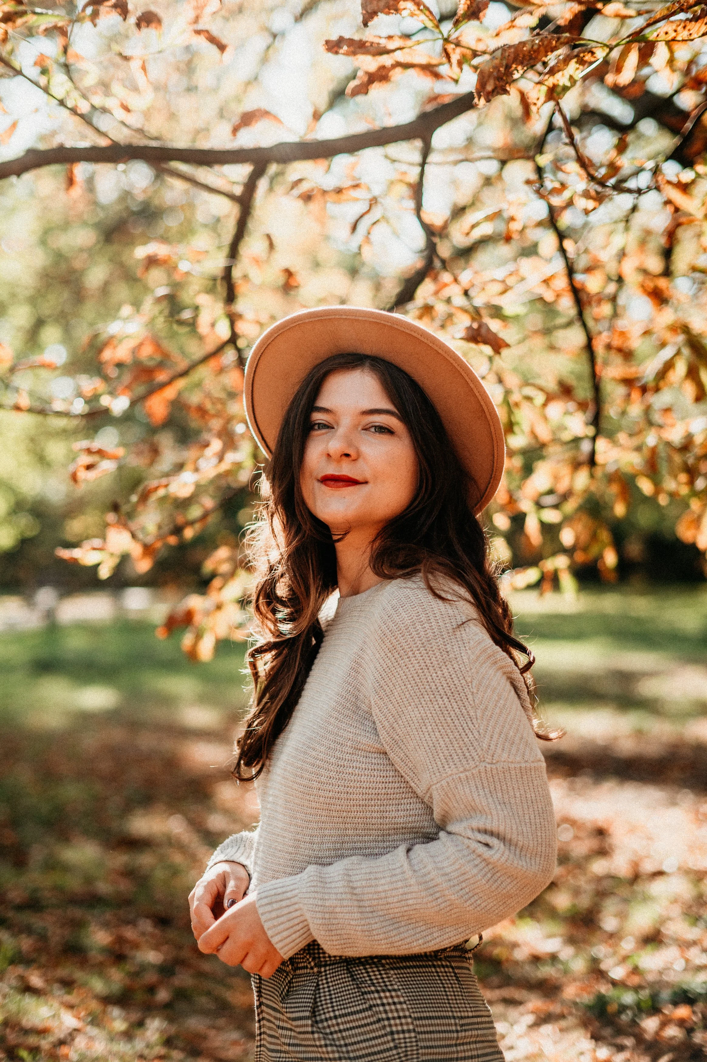 A young woman with long brown hair wearing a beige sweater, a wide-brimmed hat, and checked pants, standing outdoors in a park during autumn with colorful leaves on the tree behind her.