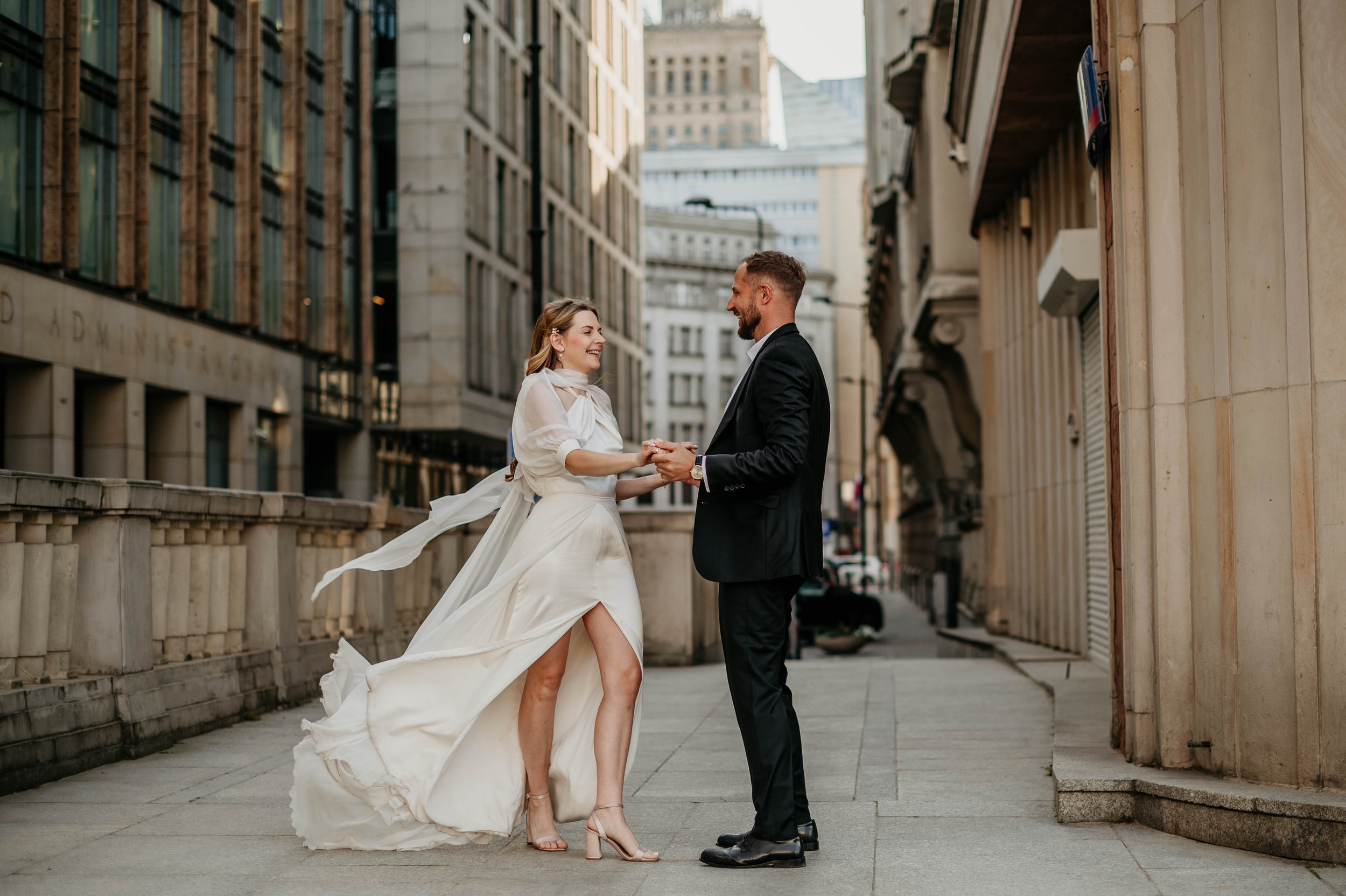 A bride and groom hold hands and smile at each other in an urban city street, with tall buildings on either side, during daytime.