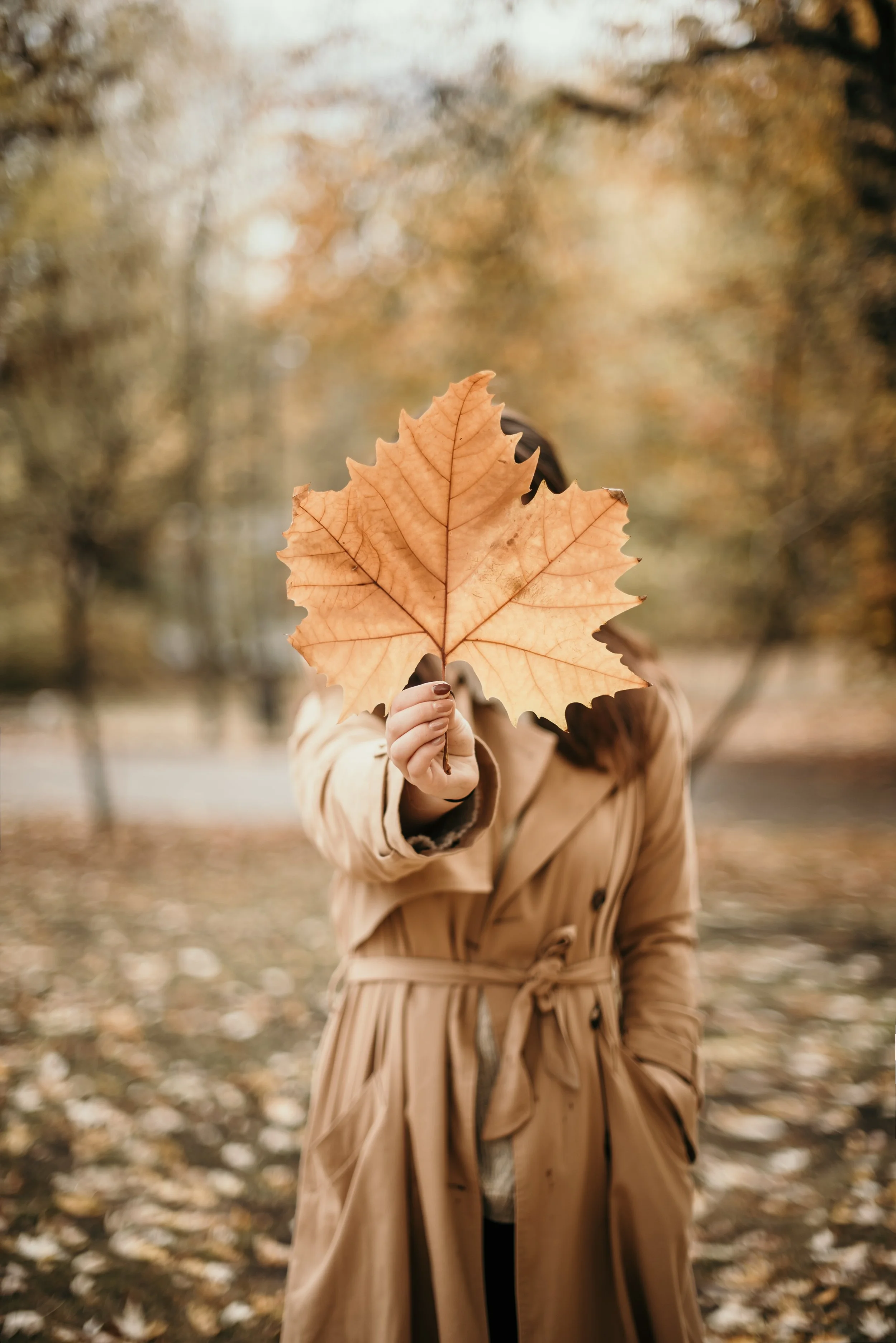 A person in a tan trench coat holding an orange autumn leaf in front of their face, standing in a park during fall with trees and fallen leaves in the background.