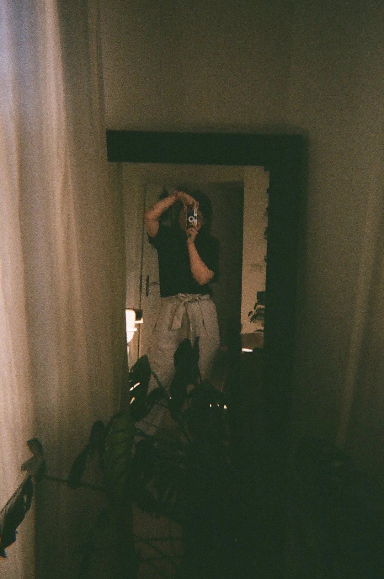 Person taking a mirror selfie with a camera in a dimly lit room, wearing a black shirt and light-colored pants with a tied shirt around the waist.