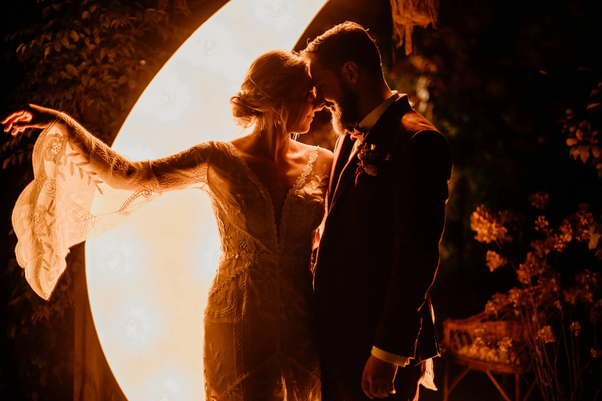 A bride and groom share an intimate moment at night, standing close with their foreheads touching, backlit by a large, warm-toned circular light, outdoors with foliage and a chair in the background.