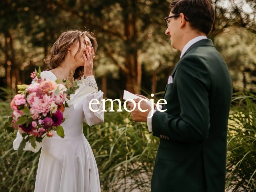 A woman in a white dress holding a bouquet of pink flowers, covering her face with her hands; a man in a suit is holding a note, facing her, in an outdoor setting with trees.