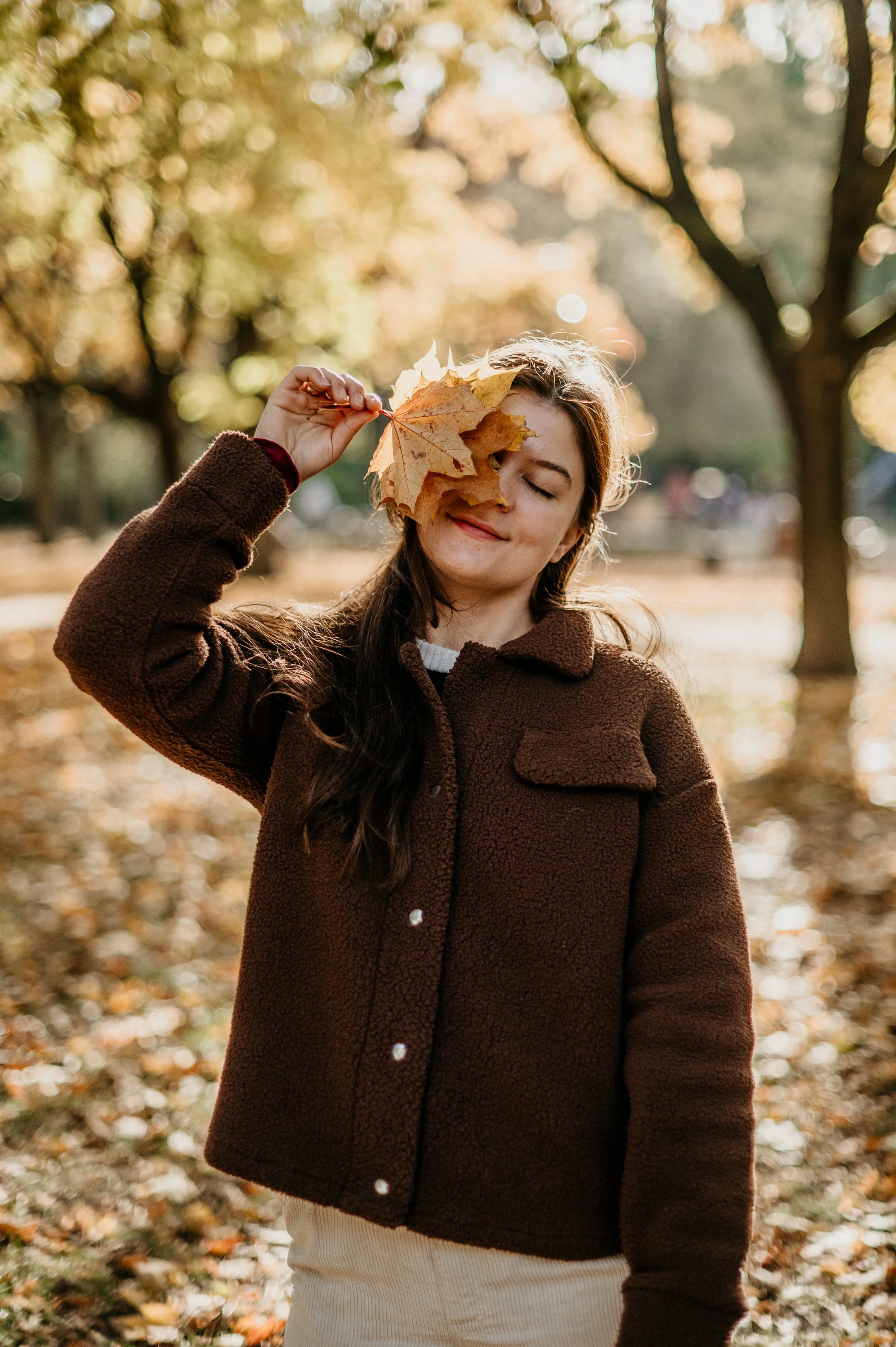 A woman in a brown coat holding autumn leaves in front of her face in a park during fall.