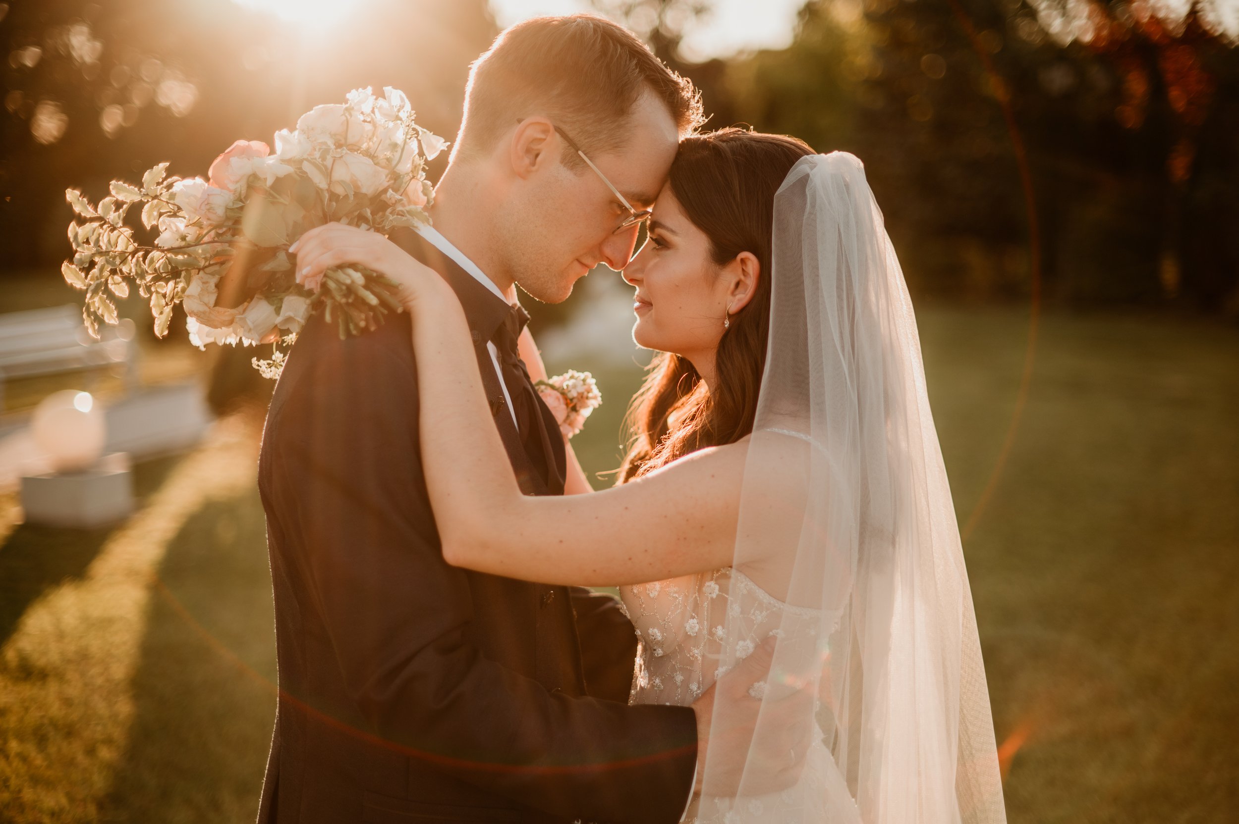 A bride and groom embrace outdoors during sunset, with their foreheads touching, holding a bouquet of flowers, and smiling.