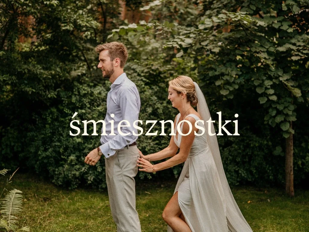 A woman in a wedding dress pulling a man in dress shirt and pants by his belt in an outdoor garden with trees and greenery, both smiling and laughing.