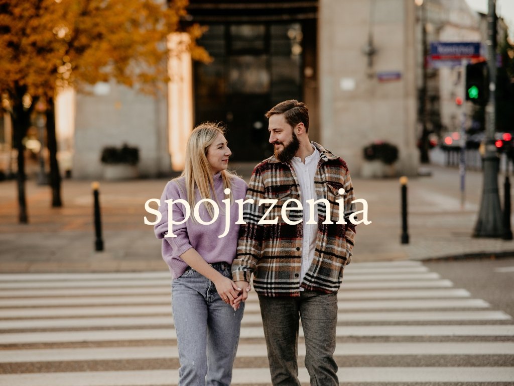 A young couple holding hands and walking across a city crosswalk during autumn, with trees and buildings in the background.