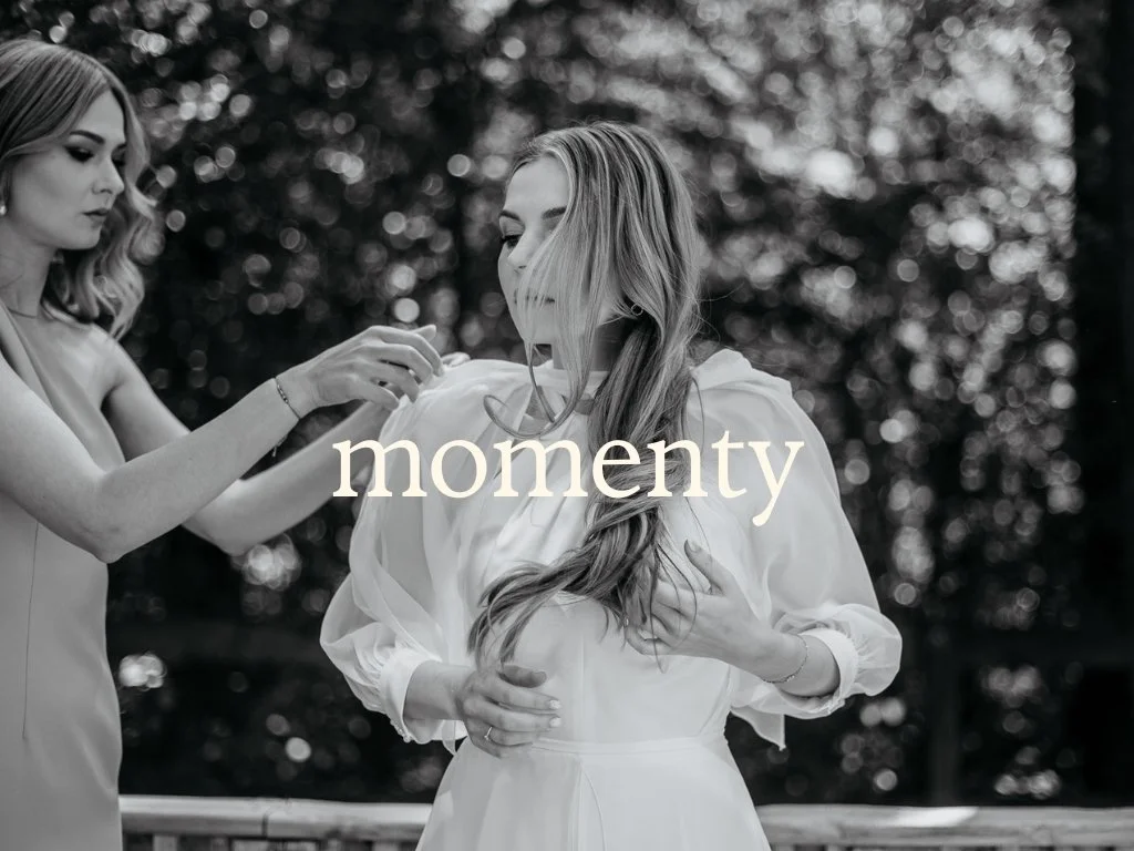A woman with long hair in a white dress is getting ready with the help of another woman in an outdoor setting, with the word 'momenty' overlaid.