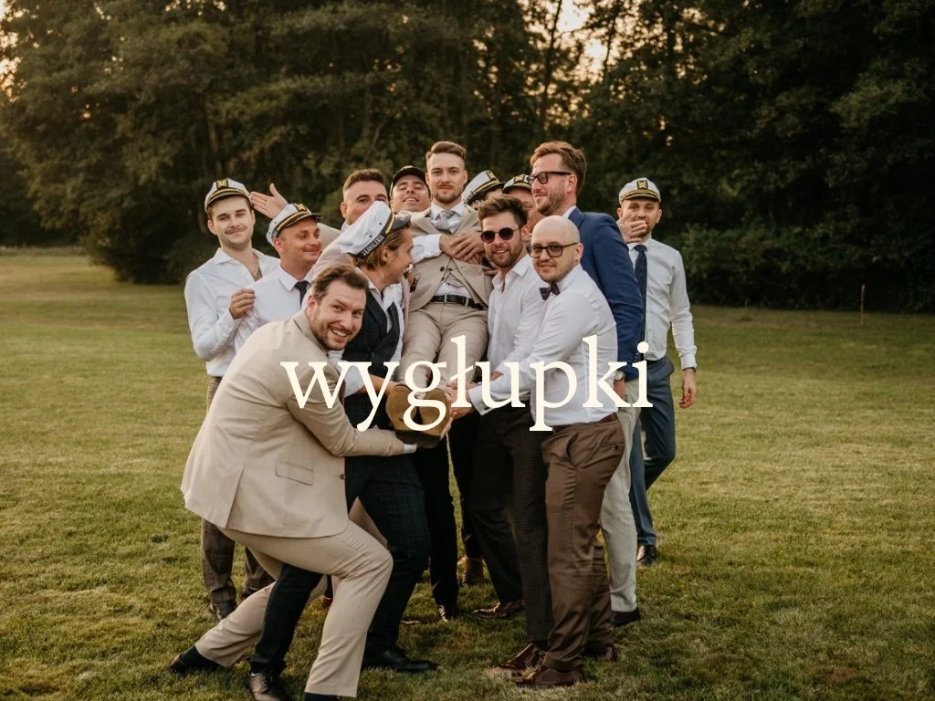 Group of men dressed in formal suits and ship captain hats, lifting a man in a suit outdoors on a grassy field with trees in the background, celebrating.