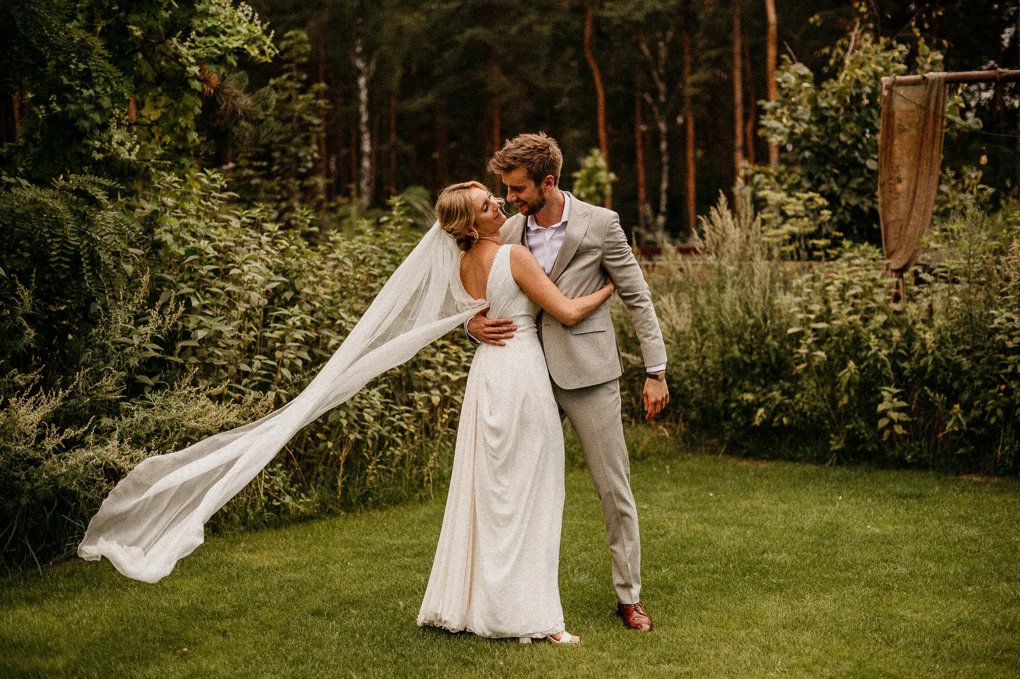 A bride in a white wedding dress and veil and a groom in a light gray suit are embracing and dancing on a grassy area outdoors, surrounded by greenery and trees.