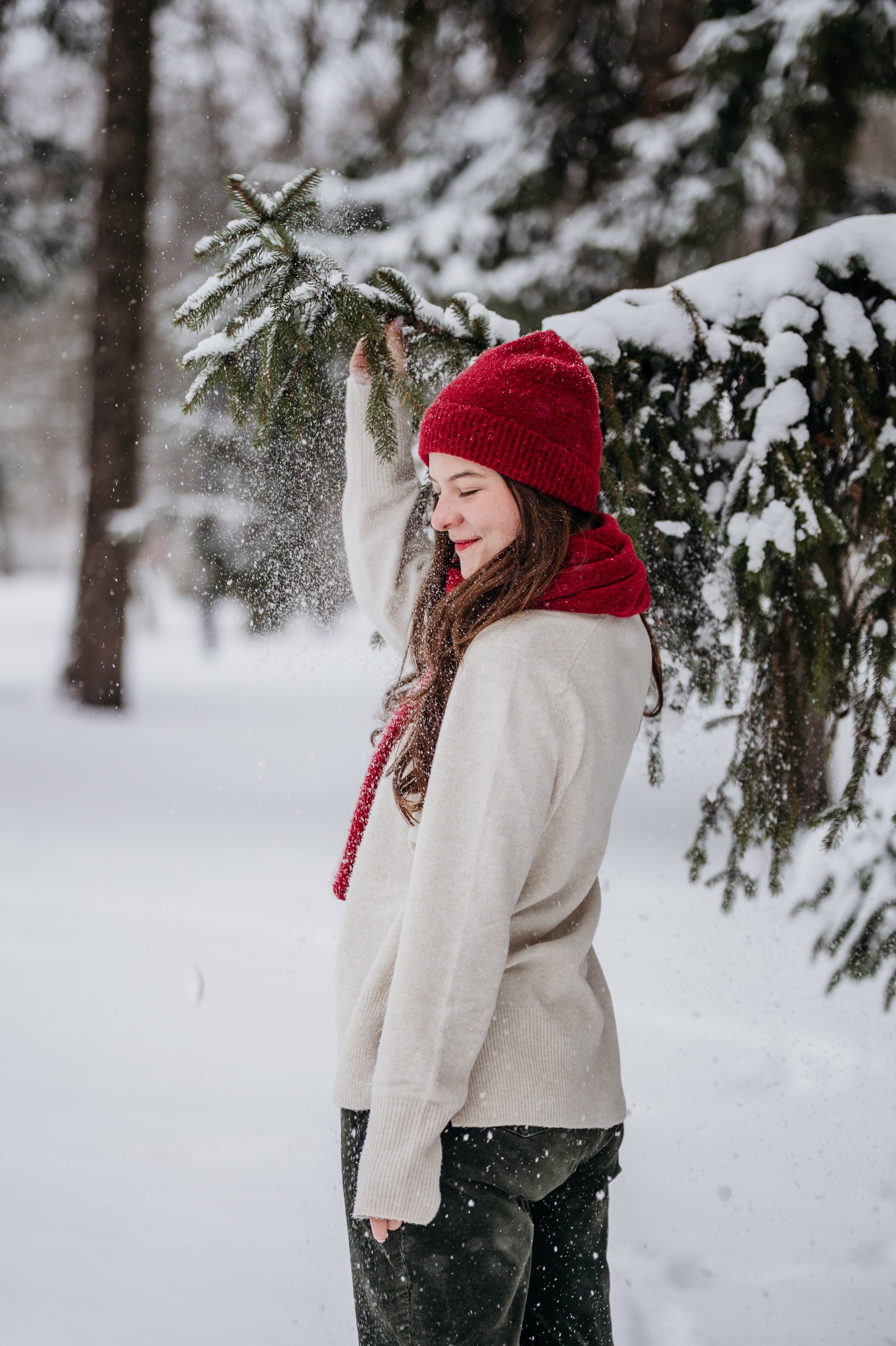 A young woman wearing a red knit hat and beige sweater smiling as she holds a branch of a snow-covered pine tree in a snowy forest.
