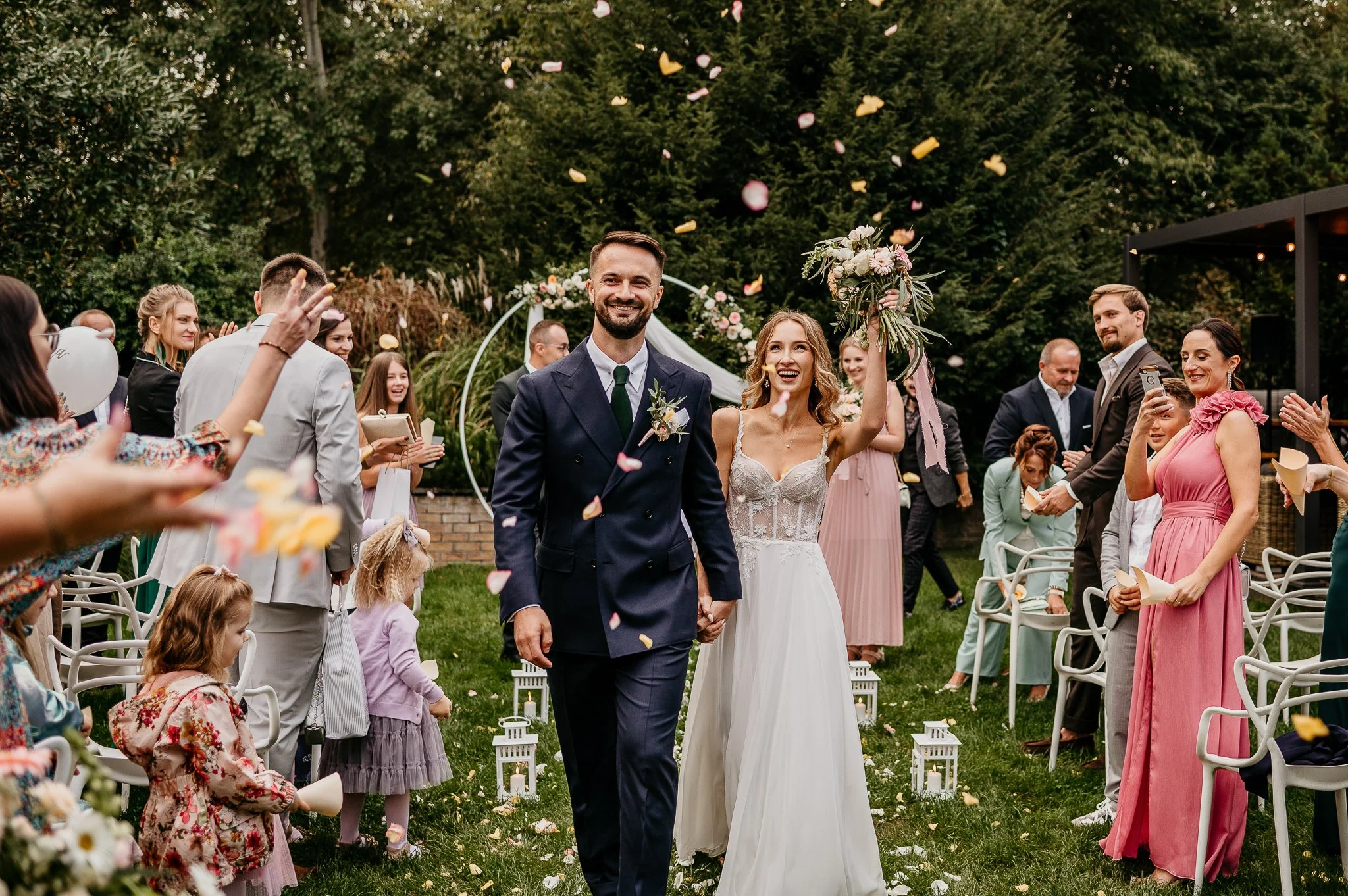 A newlywed couple, the groom in a navy suit and the bride in a white lace wedding dress, walking down the aisle holding hands, with the bride raising her bouquet in celebration. Guests around them are cheering and throwing flower petals in an outdoor garden setting with green trees and lanterns.