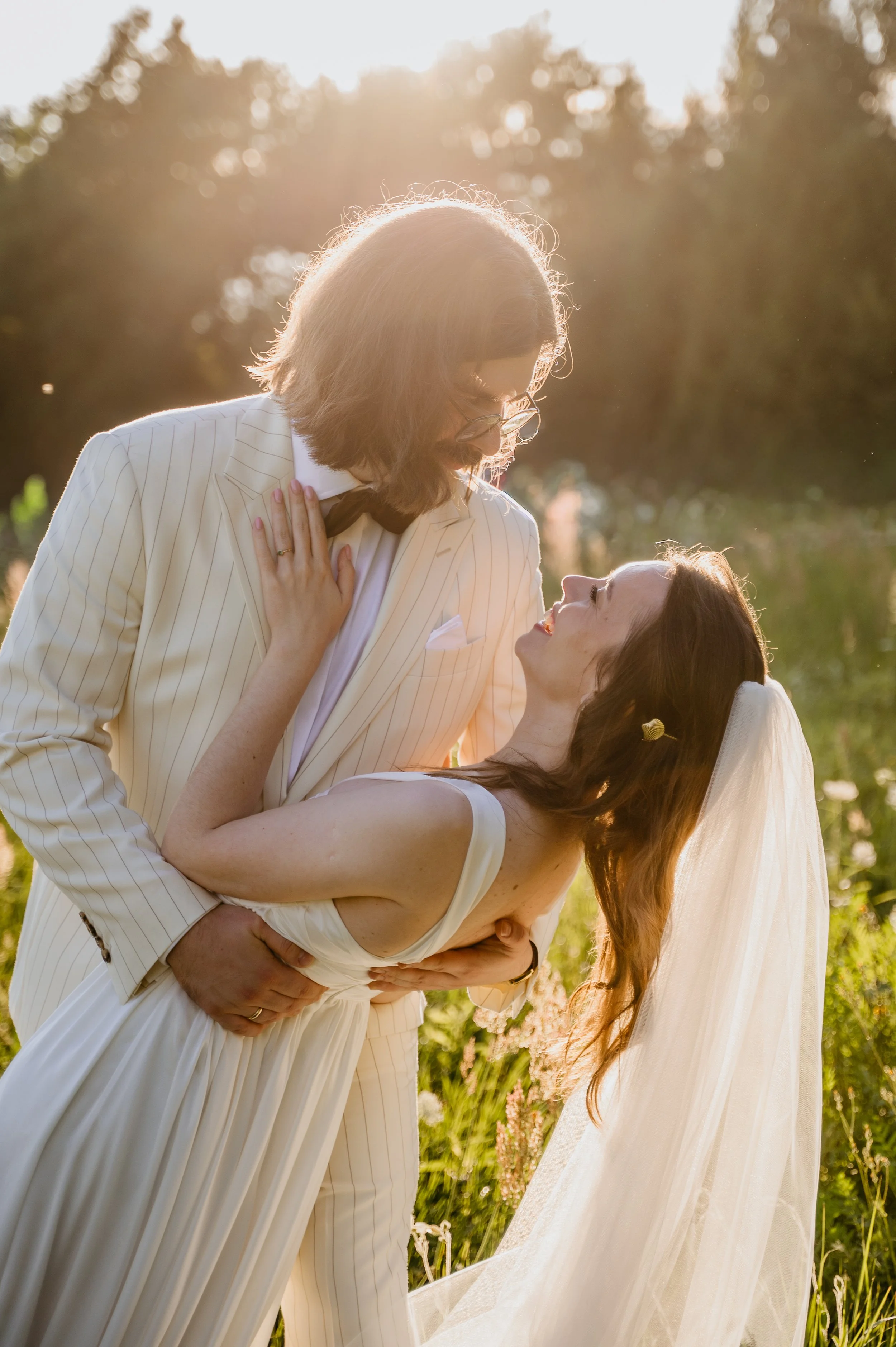 A bride and groom embrace outdoors during sunset, with the groom in a white pinstripe suit and the bride in a white wedding dress and veil.