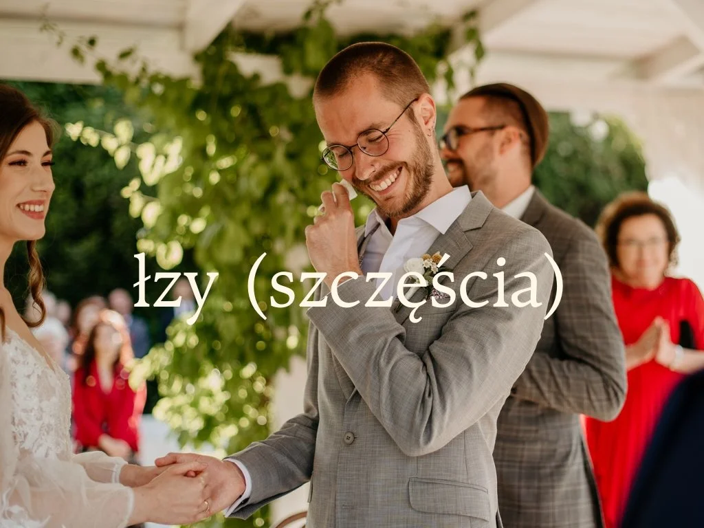 A groom in a gray suit is smiling while holding hands with a bride during their wedding ceremony, surrounded by guests.