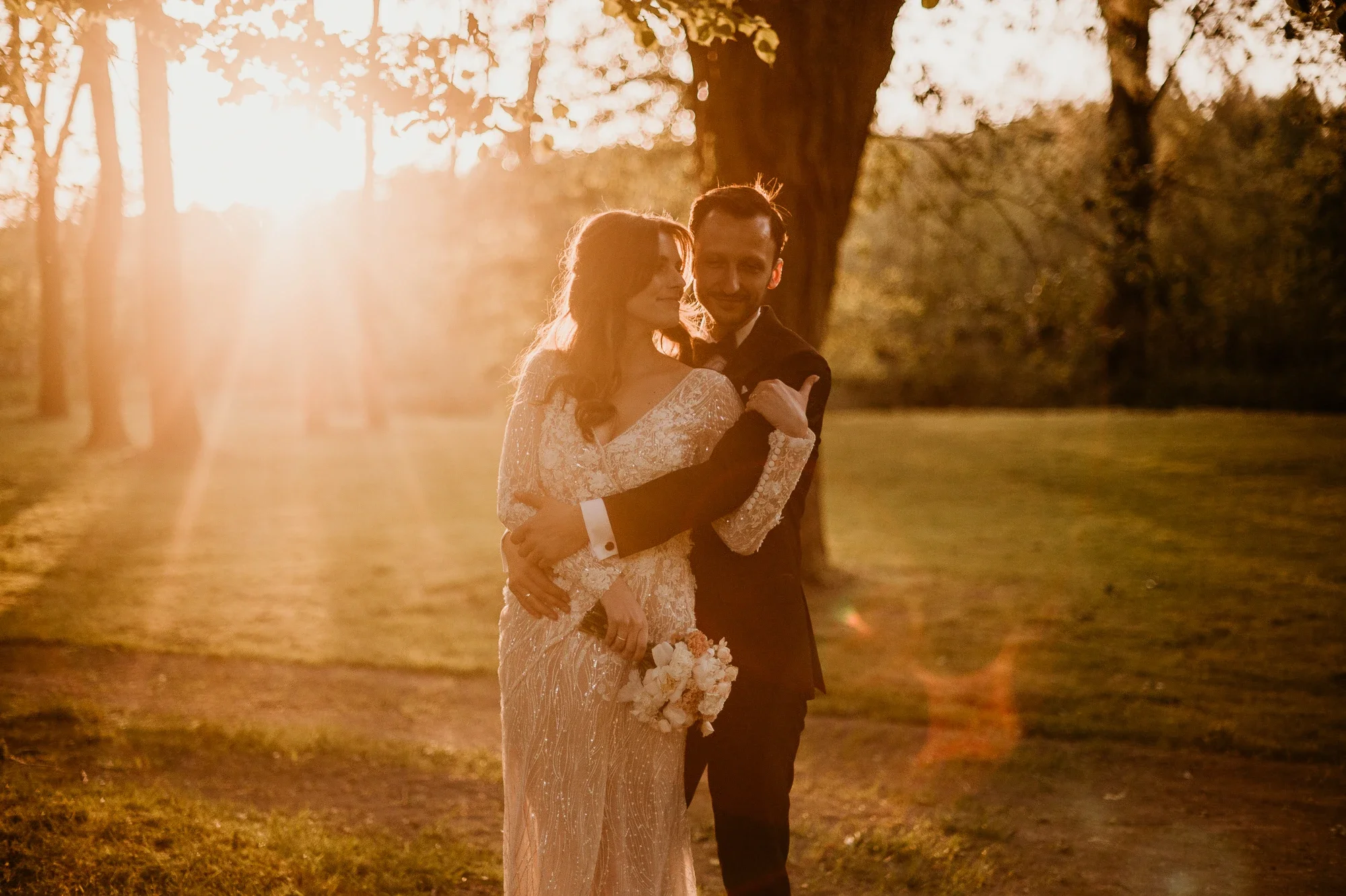 A bride and groom hugging outdoors during sunset, the bride holding a bouquet of flowers, surrounded by trees and open field.