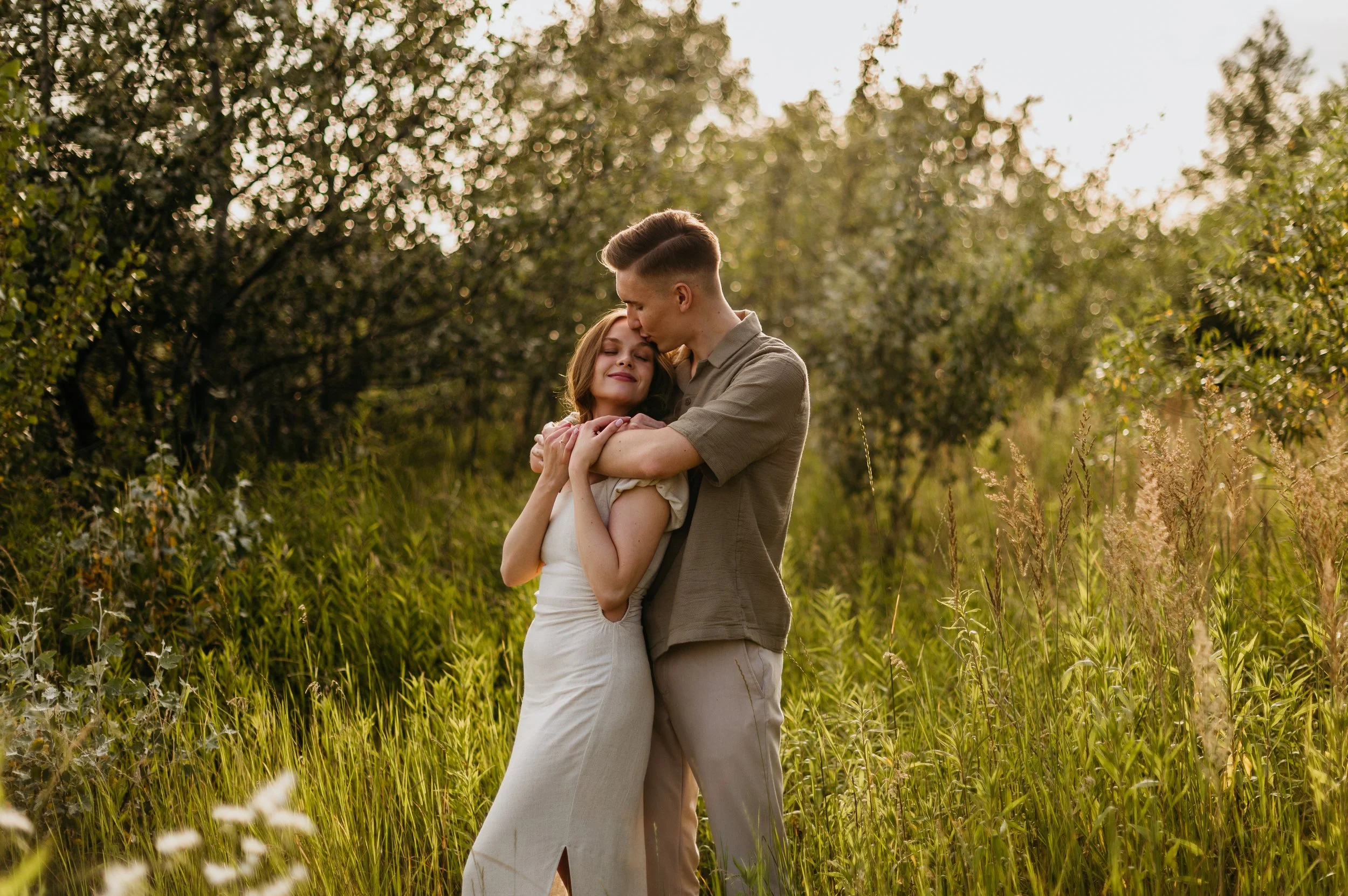 A young couple sharing a tender moment outdoors near sunset, with trees and tall grass in the background.