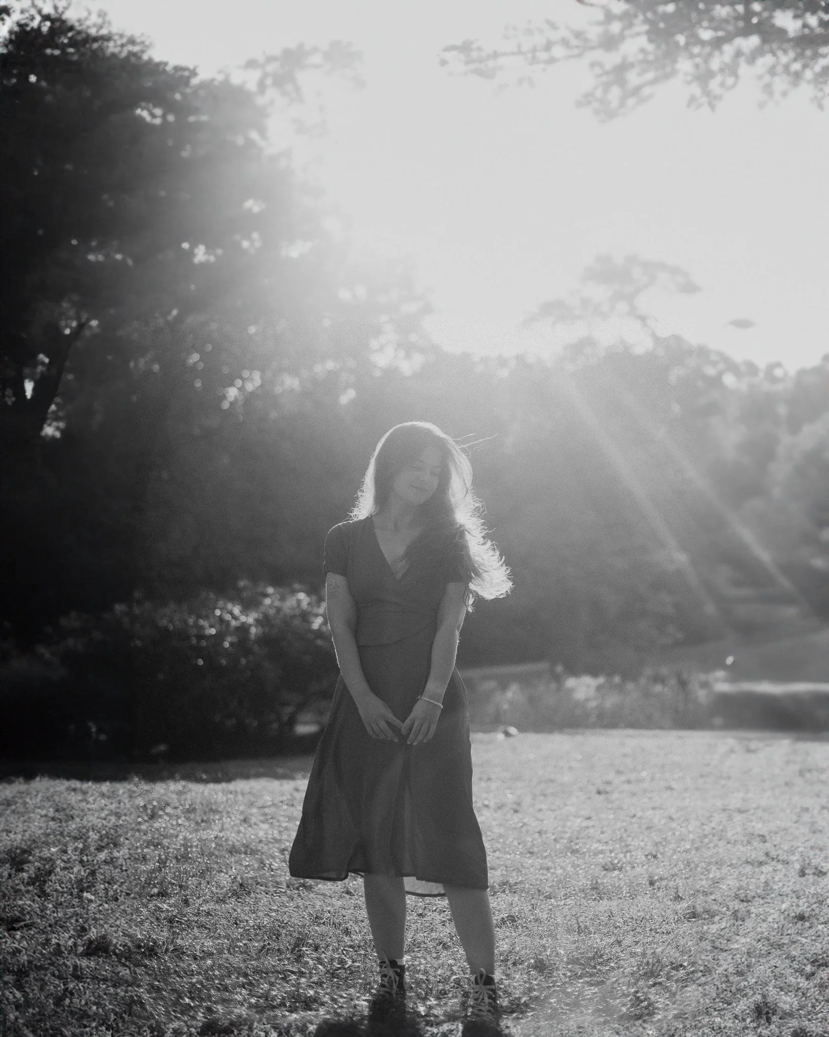 A woman with long hair standing outdoors in a grassy area, with sunlight shining brightly behind her, creating a silhouette effect in a black and white photo.