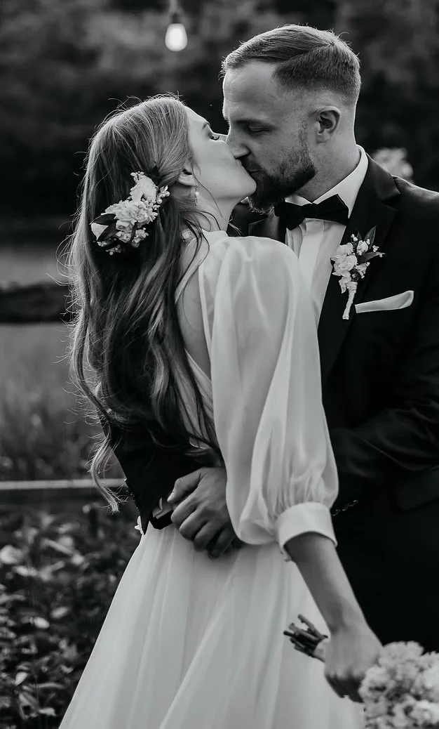 A black-and-white photo of a bride and groom sharing a kiss, outdoors with a blurred background and a hanging light bulb above them. The bride is wearing a white dress with long sleeves and has a floral hairpiece. The groom is in a tuxedo with a bow tie and a boutonniere.