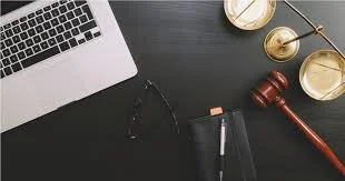 Close-up of a workspace with a laptop, glasses, a notebook, a pen, and a gavel on a dark wooden table.