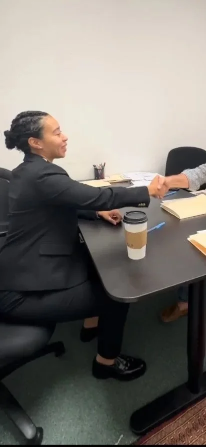 A woman paralegal in a black suit smiling and shaking hands with someone across a table in a business setting, with documents and a coffee cup on the table.