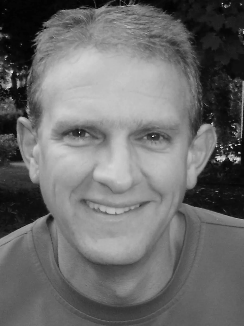 A smiling man with short, light-colored hair, looking directly at the camera, outdoors with blurred trees in the background.