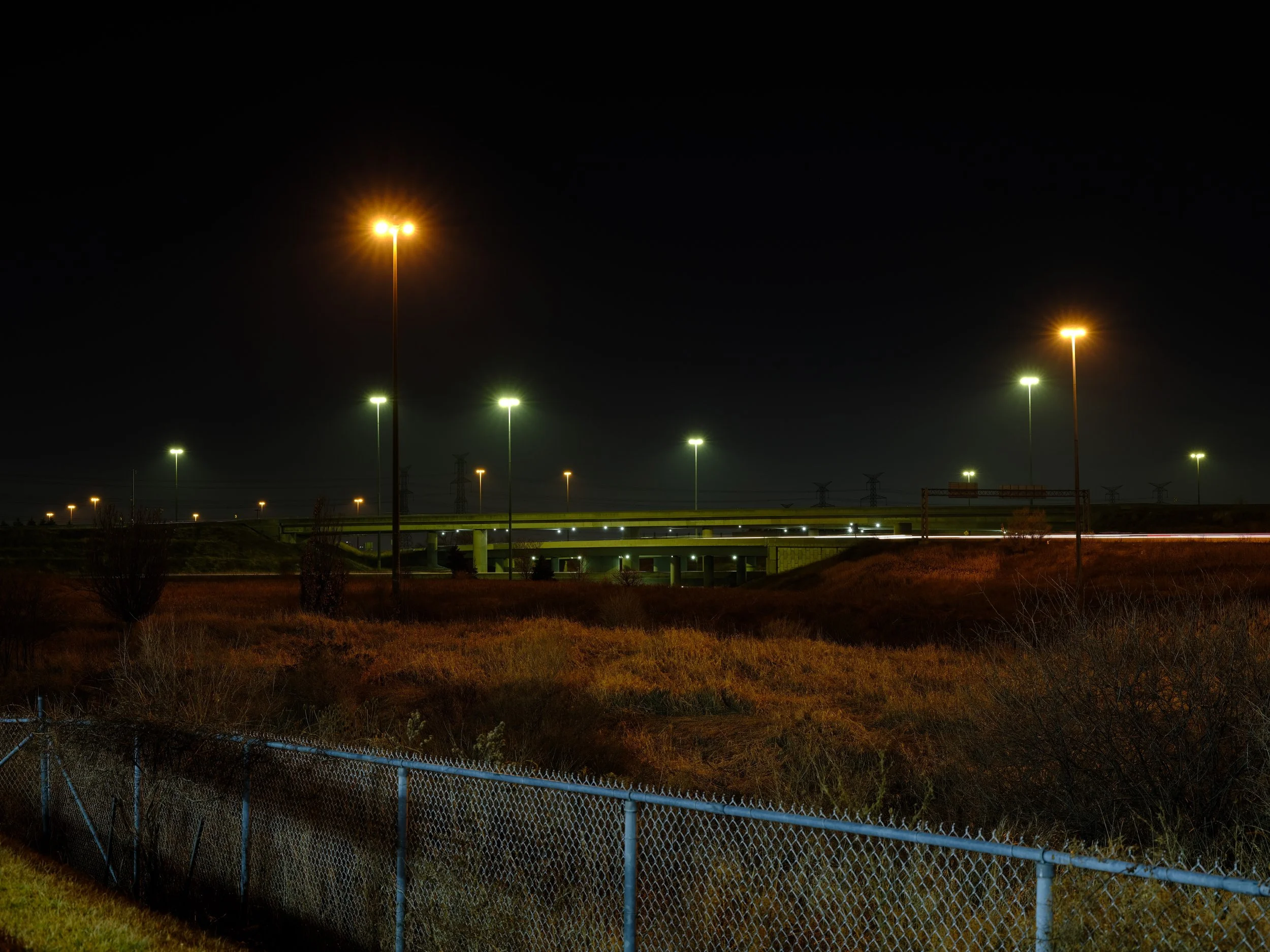 Night scene of a highway overpass illuminated by streetlights, with a chain-link fence in the foreground and power lines in the distance.