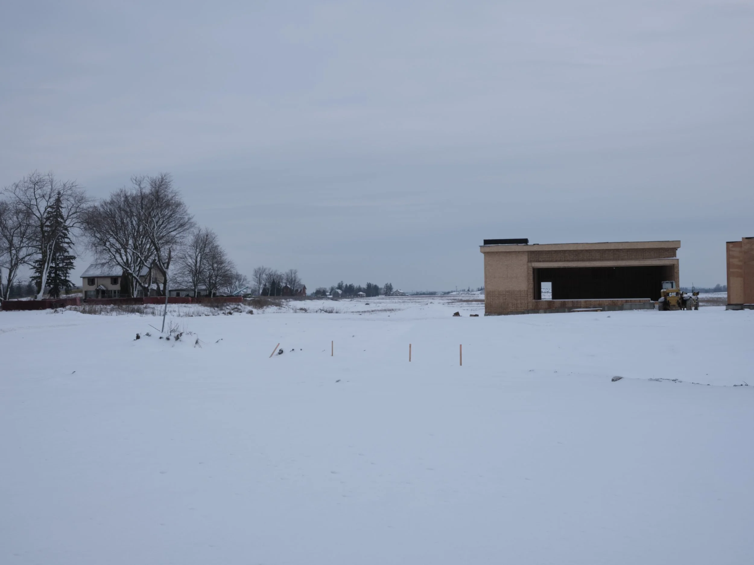 Snow-covered construction site with a partially built brick building, a small bulldozer, and leafless trees in the background.