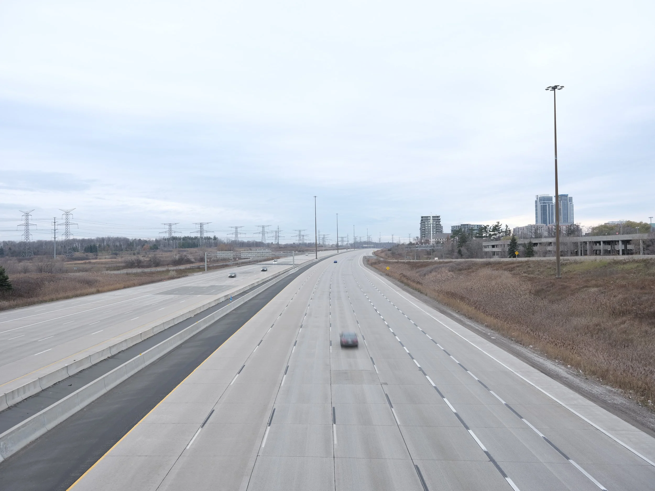 A mostly empty highway with a few moving cars, some buildings and power lines in the background, and a cloudy sky overhead.