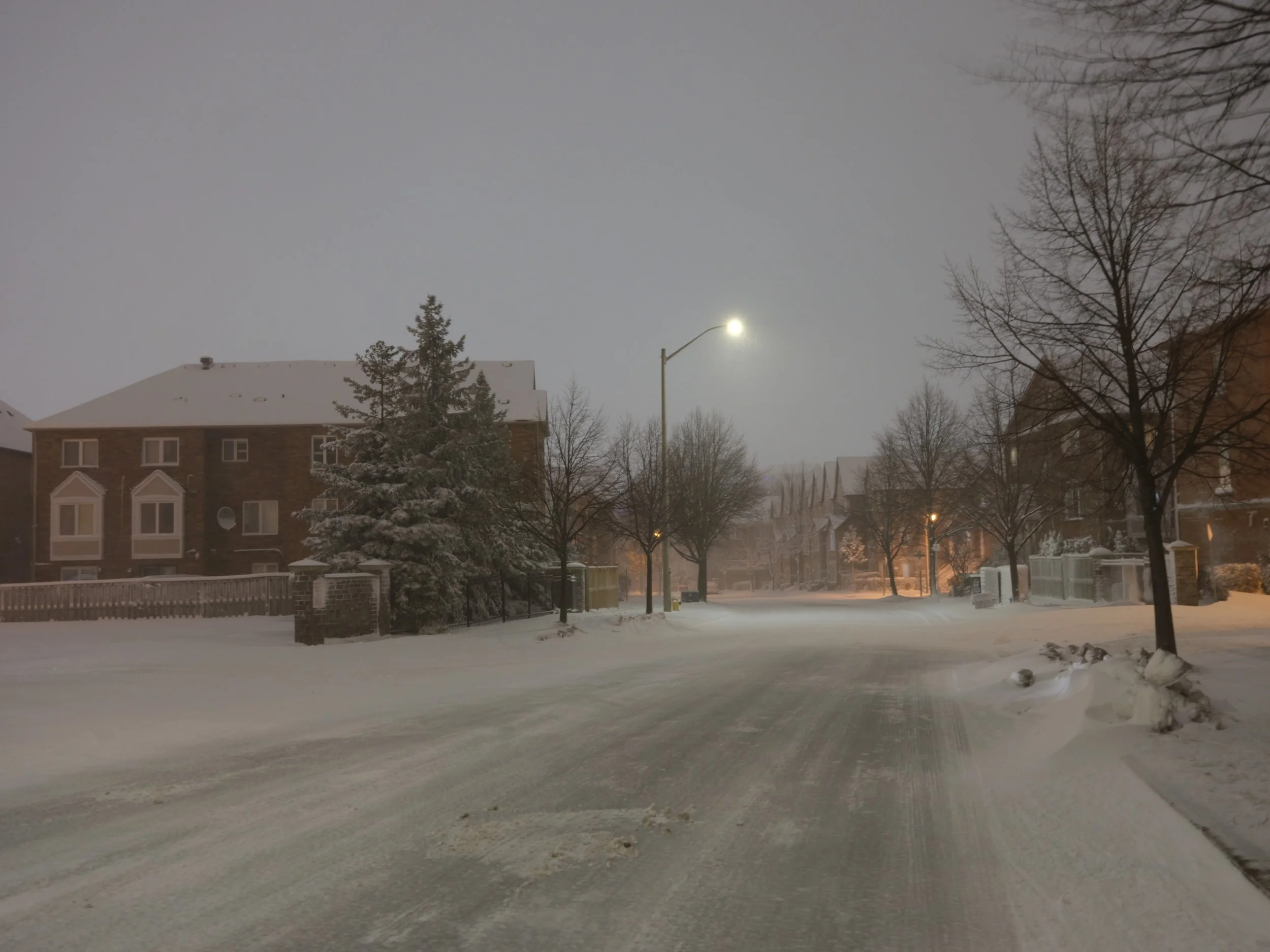 Snowy residential street at night with snow-covered houses, trees, and a streetlight illuminating the scene.