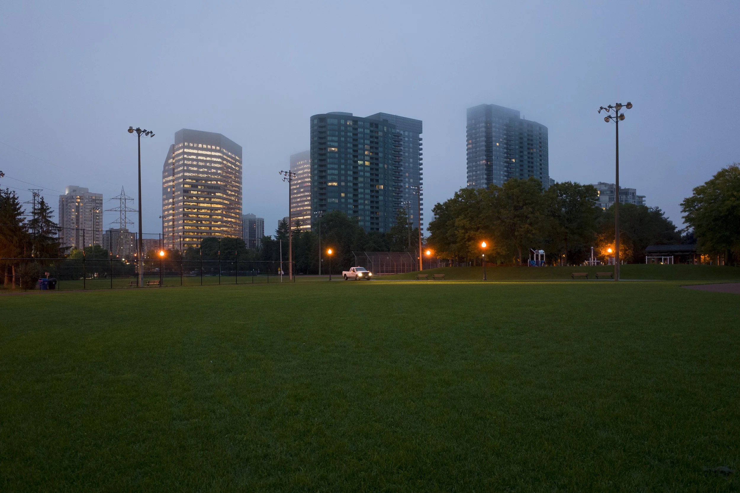 Maintenance track in a city park at dusk. Modern high-rise buildings with lit windows in the background are partially obscured by early evening fog.