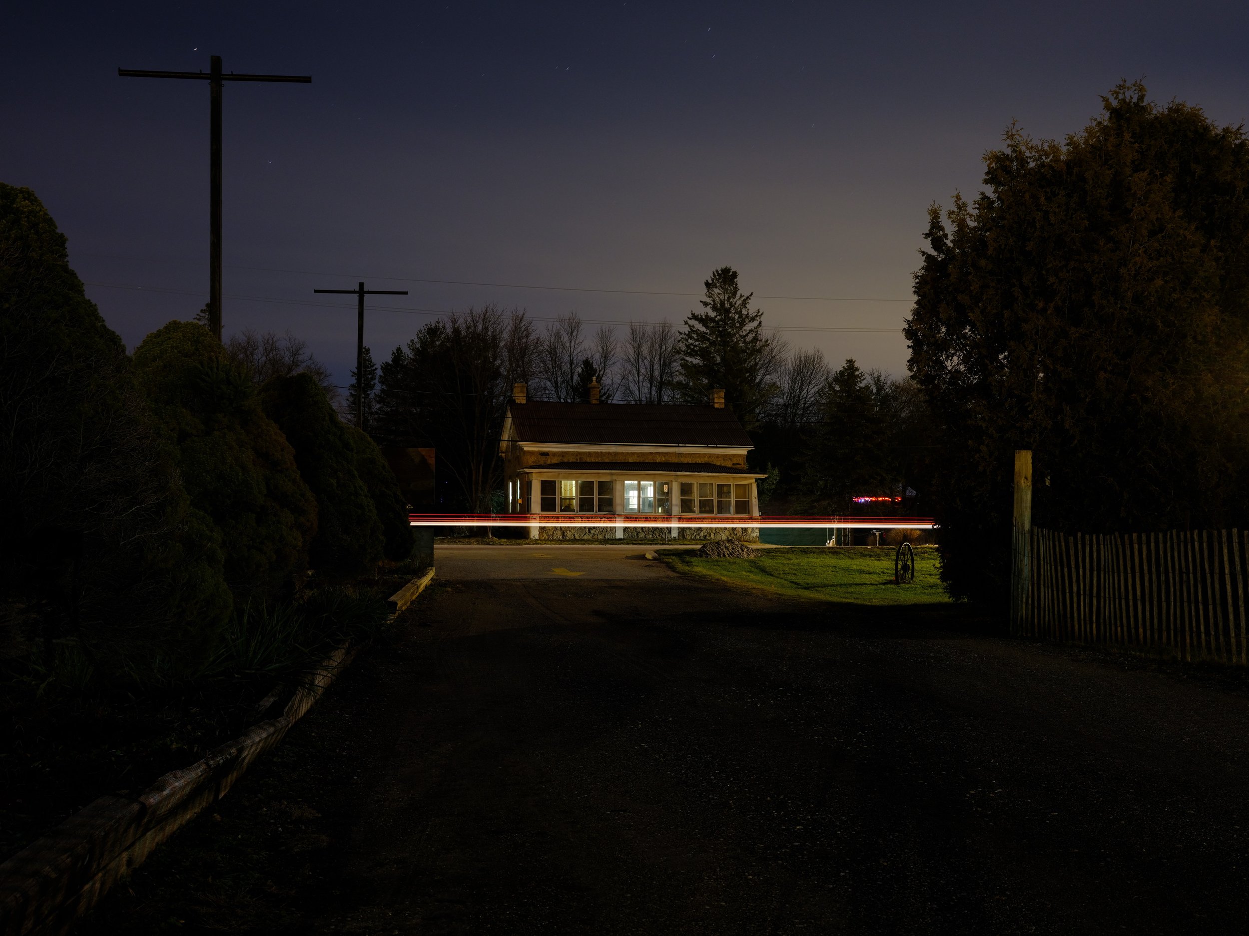 A house with lit windows at night, surrounded by trees and shrubs, with power poles and light trails from passing vehicles visible in the scene.
