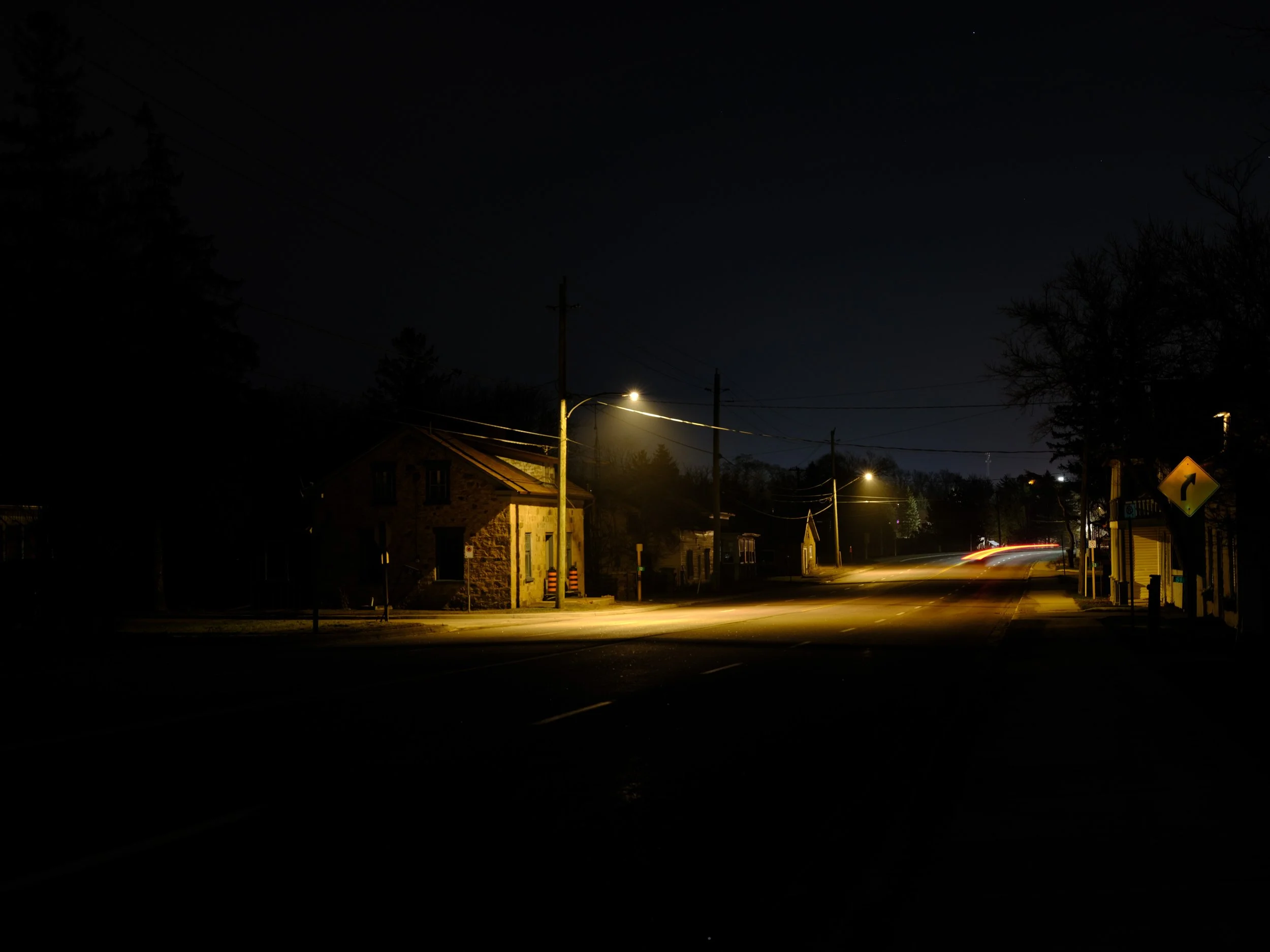 Nighttime scene of a small town street with a few lit buildings, streetlights, and trees, with some light streaks from moving vehicles.